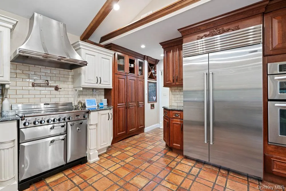 Kitchen featuring backsplash, appliances with stainless steel finishes, beam ceiling, ventilation hood, and dark stone countertops Kitchen featuring backsplash, appliances with stainless steel finishes, beam ceiling, ventilation hood, and dark stone countertops