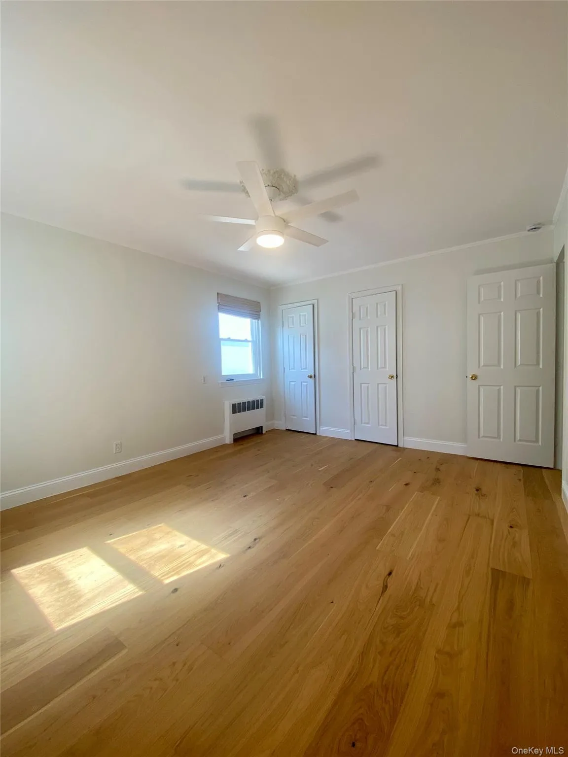 Unfurnished bedroom with two closets, light wood-style flooring, radiator, a ceiling fan, and crown molding Unfurnished bedroom with two closets, light wood-style flooring, radiator, a ceiling fan, and crown molding