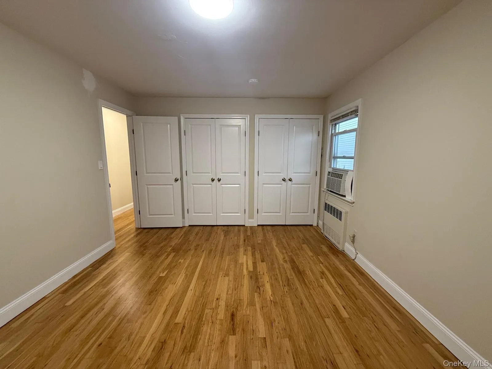Unfurnished bedroom featuring two closets, light wood-type flooring, radiator heating unit, and cooling unit Unfurnished bedroom featuring two closets, light wood-type flooring, radiator heating unit, and cooling unit