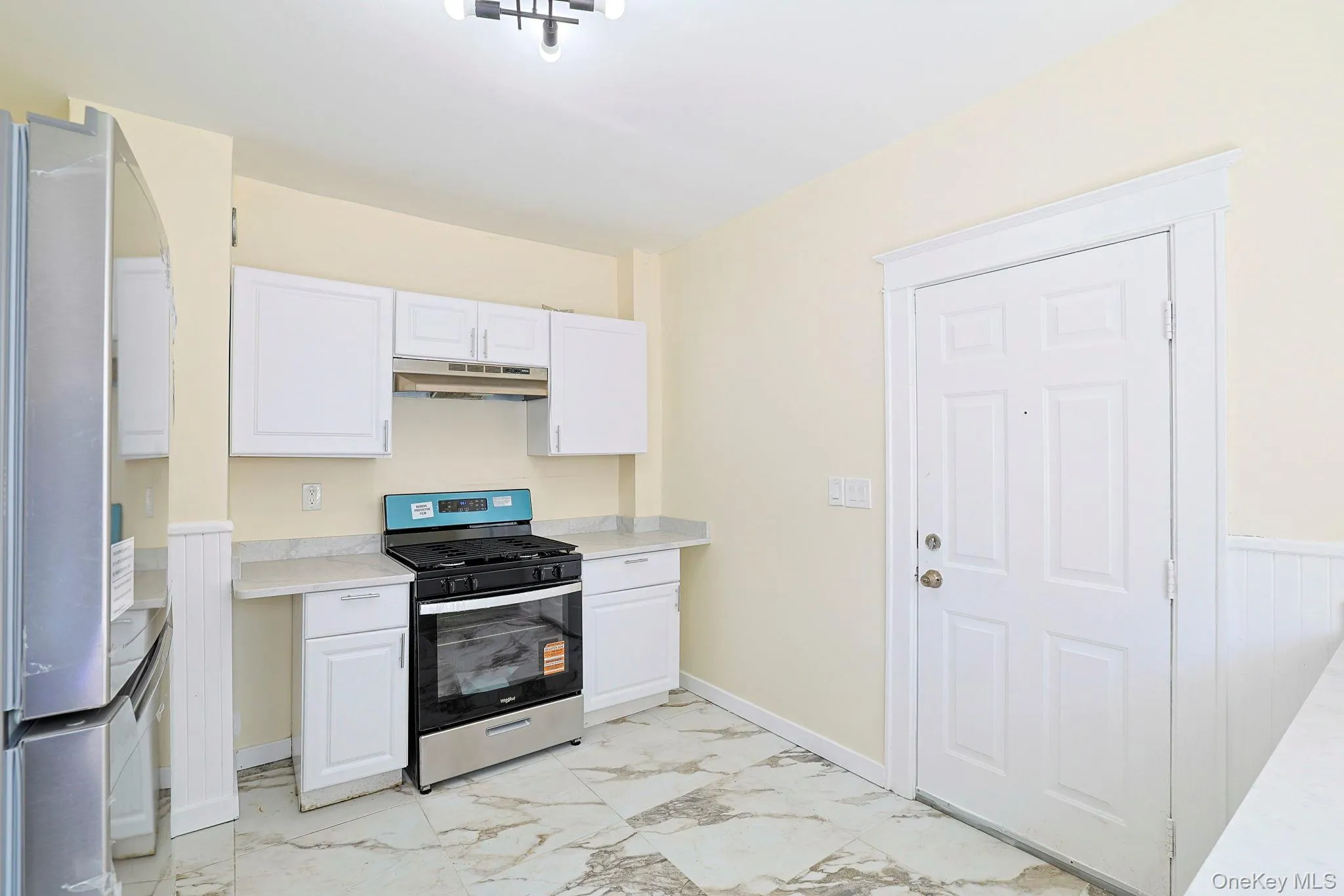 Kitchen featuring stainless steel appliances, light countertops, light marble finish floors, white cabinetry, and under cabinet range hood Kitchen featuring stainless steel appliances, light countertops, light marble finish floors, white cabinetry, and under cabinet range hood