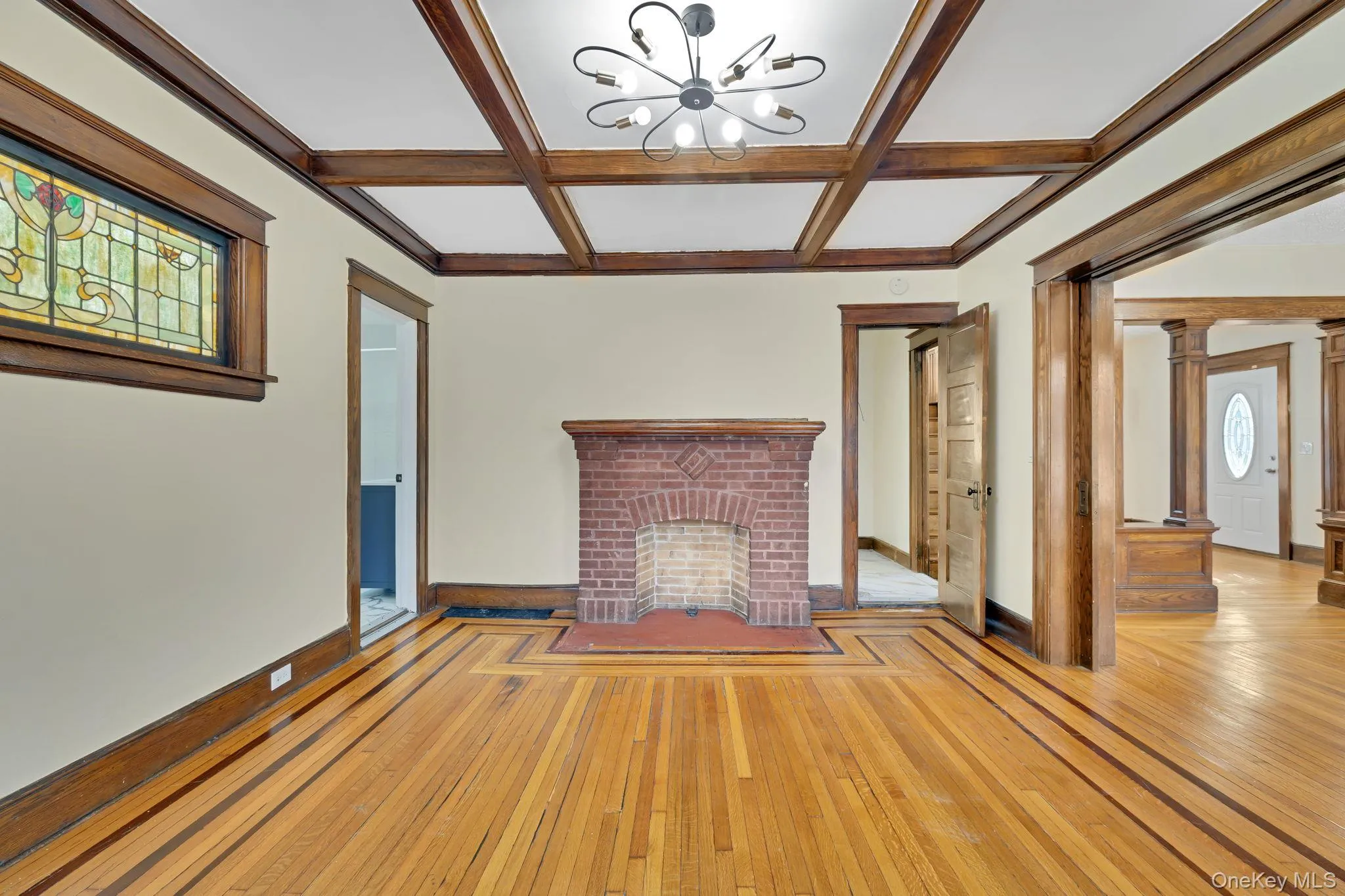Unfurnished living room featuring hardwood / wood-style flooring, coffered ceiling, beamed ceiling, a brick fireplace, and a chandelier Unfurnished living room featuring hardwood / wood-style flooring, coffered ceiling, beamed ceiling, a brick fireplace, and a chandelier