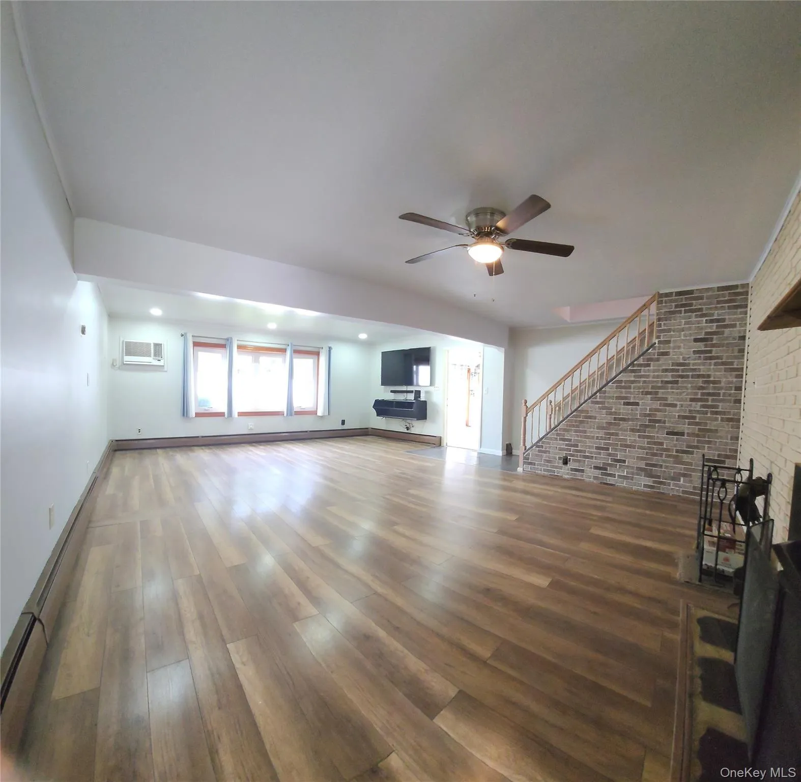 Unfurnished living room with baseboard heating, brick wall, wood finished floors, a ceiling fan, and a wall mounted AC Unfurnished living room with baseboard heating, brick wall, wood finished floors, a ceiling fan, and a wall mounted AC