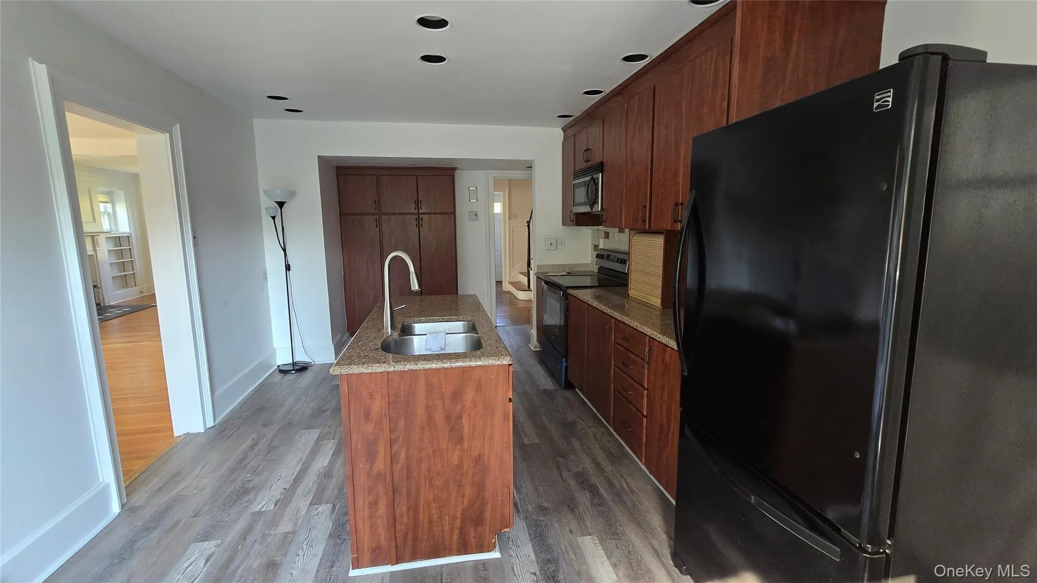 Kitchen with black appliances, dark wood-style flooring, an island with sink, light stone countertops, and brown cabinetry Kitchen with black appliances, dark wood-style flooring, an island with sink, light stone countertops, and brown cabinetry