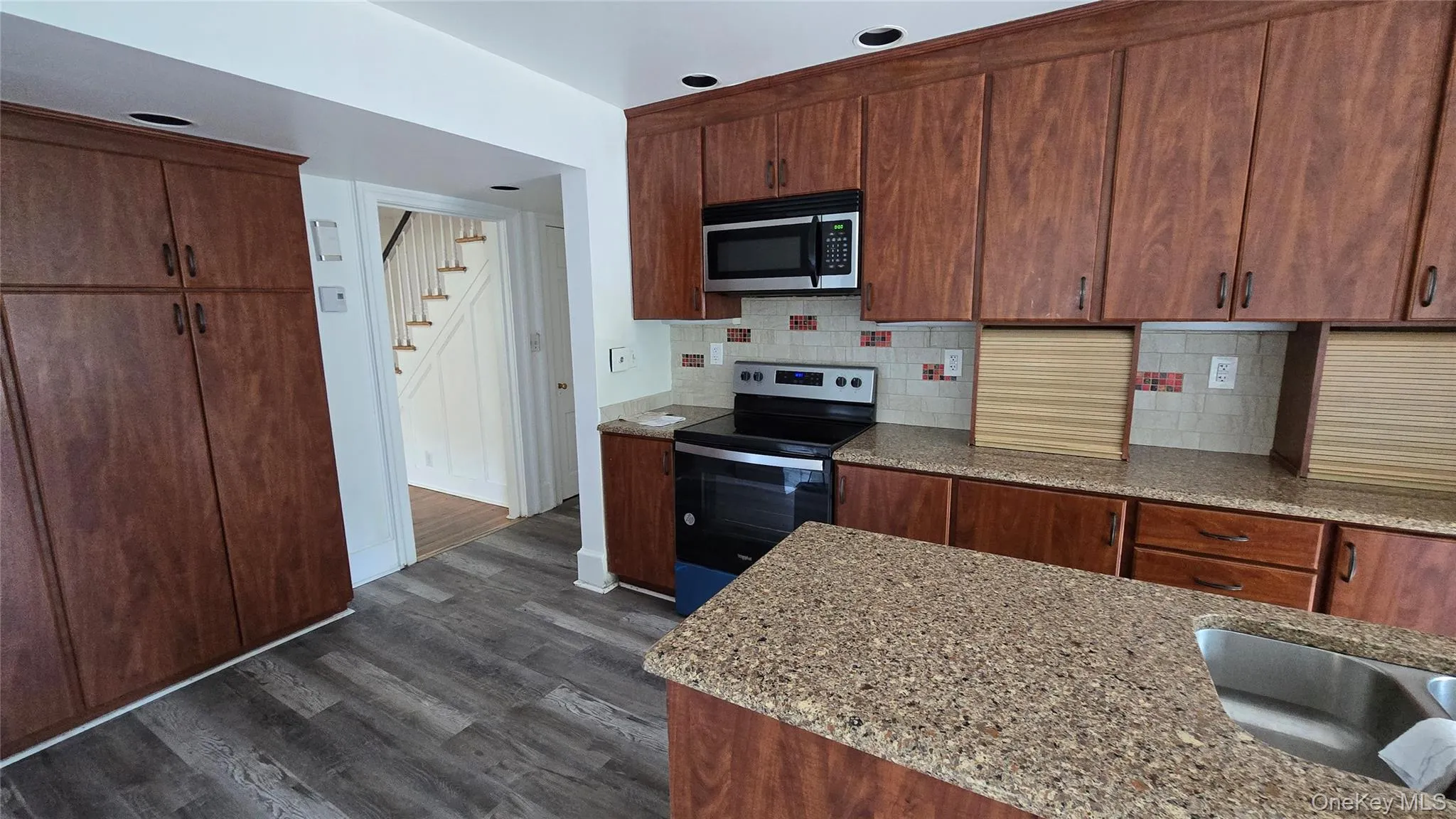 Kitchen featuring stainless steel appliances, dark wood-type flooring, decorative backsplash, light stone countertops, and brown cabinetry Kitchen featuring stainless steel appliances, dark wood-type flooring, decorative backsplash, light stone countertops, and brown cabinetry