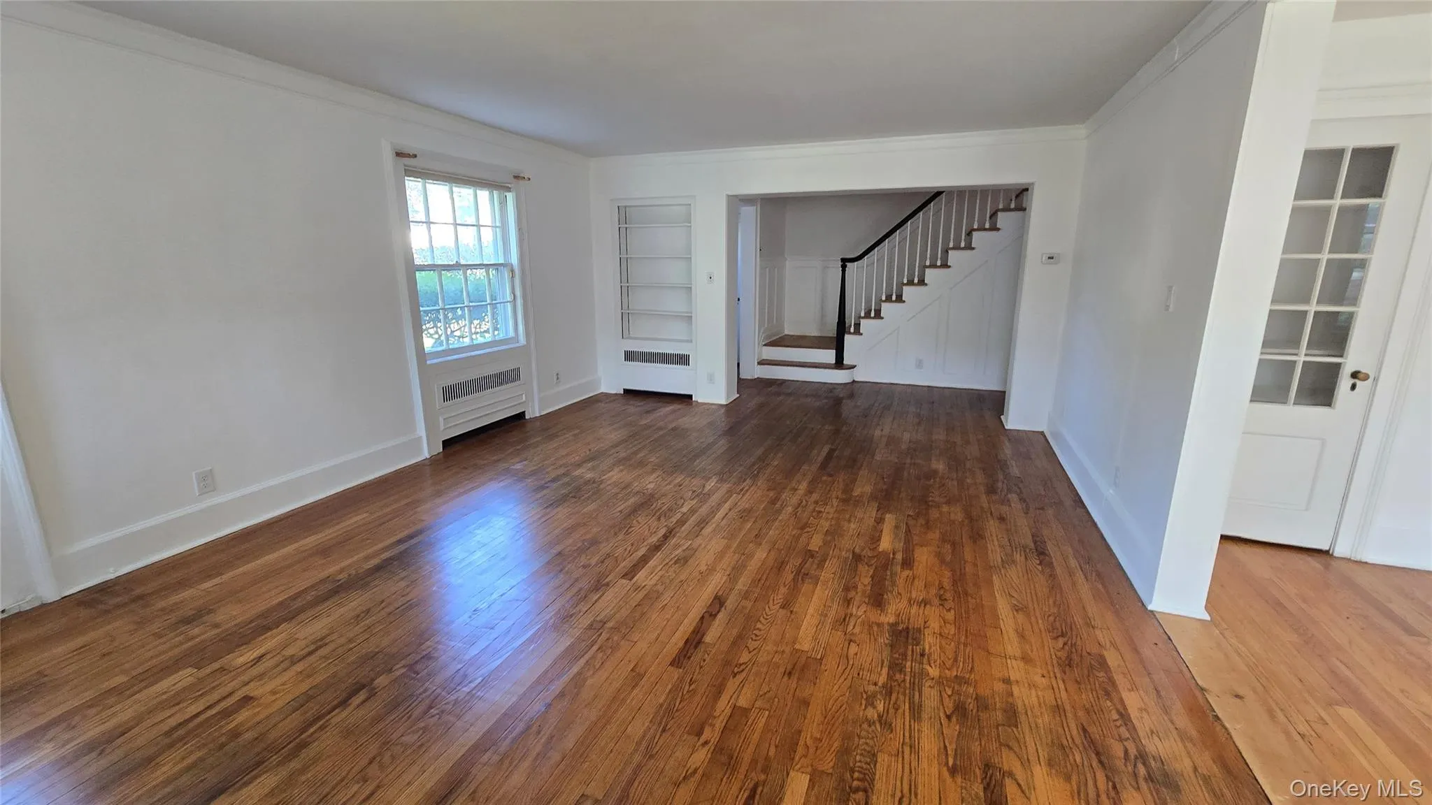 Unfurnished living room featuring ornamental molding, dark wood-style floors, built in features, stairs, and radiator heating unit Unfurnished living room featuring ornamental molding, dark wood-style floors, built in features, stairs, and radiator heating unit