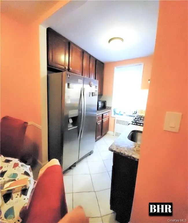 Kitchen featuring stainless steel fridge with ice dispenser, light stone countertops, light tile patterned flooring, radiator, and black microwave Kitchen featuring stainless steel fridge with ice dispenser, light stone countertops, light tile patterned flooring, radiator, and black microwave