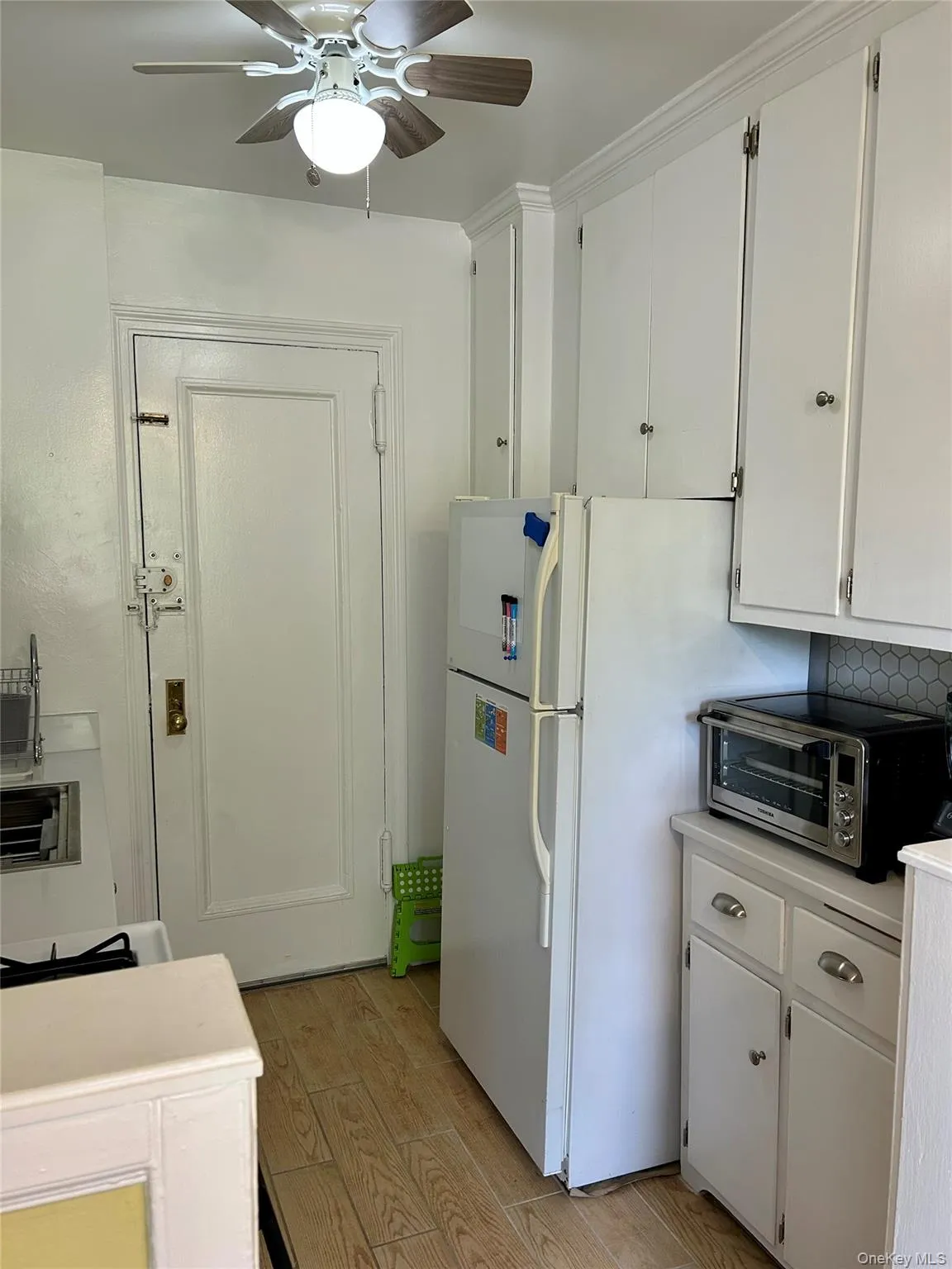 Kitchen with white cabinetry, light tile flooring, freestanding refrigerator, light countertops, and ceiling fan. Door to hallway. Kitchen with white cabinetry, light tile flooring, freestanding refrigerator, light countertops, and ceiling fan. Door to hallway.