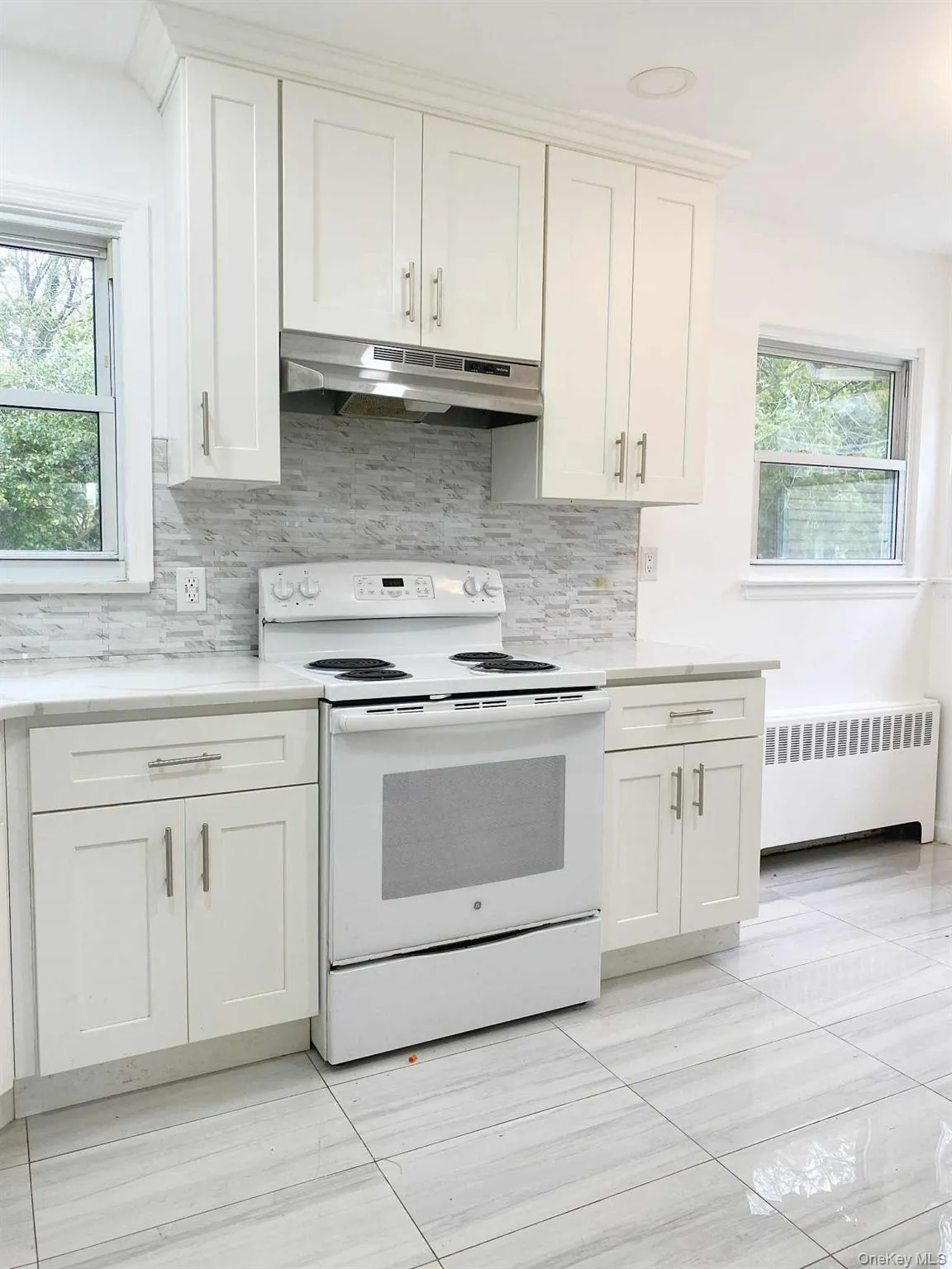 Kitchen featuring white electric stove, white cabinetry, radiator heating unit, under cabinet range hood, and backsplash Kitchen featuring white electric stove, white cabinetry, radiator heating unit, under cabinet range hood, and backsplash