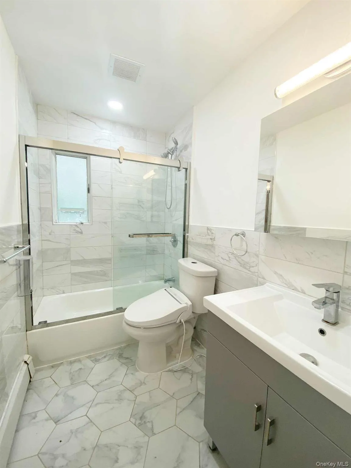 Bathroom with tile walls, a wainscoted wall, vanity, bath / shower combo with glass door, and light marble finish floors Bathroom with tile walls, a wainscoted wall, vanity, bath / shower combo with glass door, and light marble finish floors