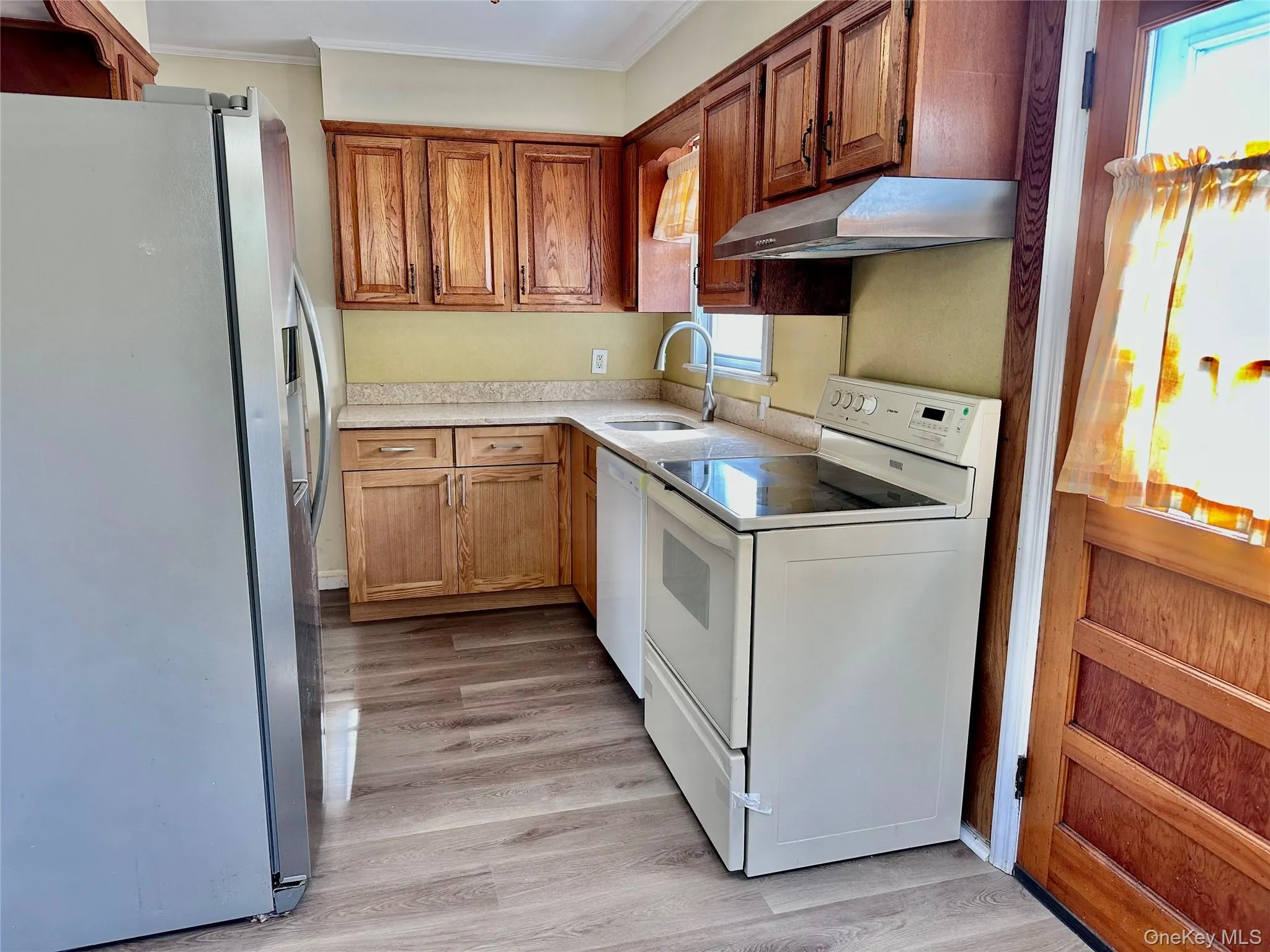 Kitchen with white appliances, light countertops, brown cabinetry, light wood finished floors, and under cabinet range hood Kitchen with white appliances, light countertops, brown cabinetry, light wood finished floors, and under cabinet range hood