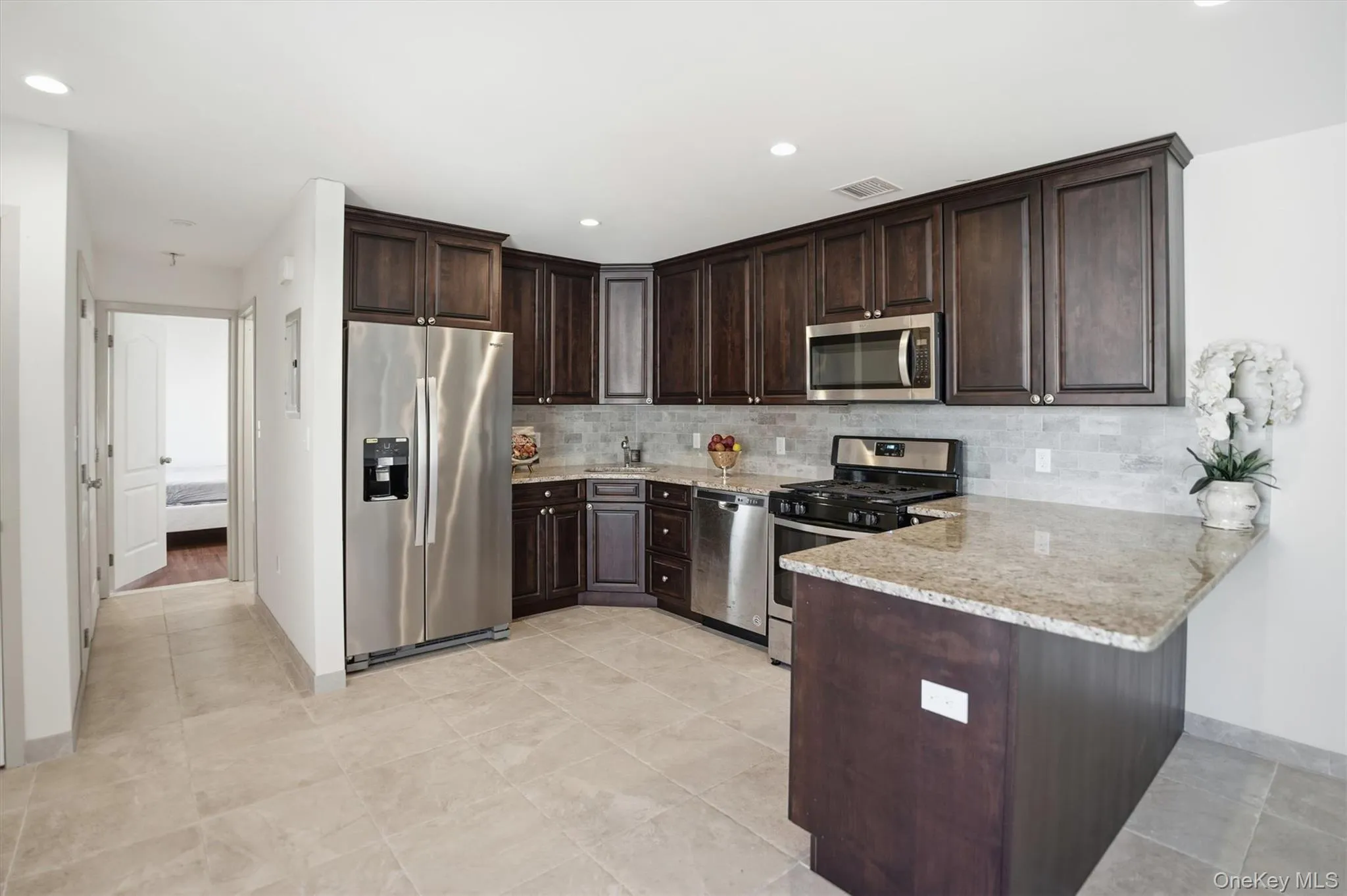 Kitchen with dark brown cabinetry, stainless steel appliances, tasteful backsplash, light stone counters, and a peninsula Kitchen with dark brown cabinetry, stainless steel appliances, tasteful backsplash, light stone counters, and a peninsula
