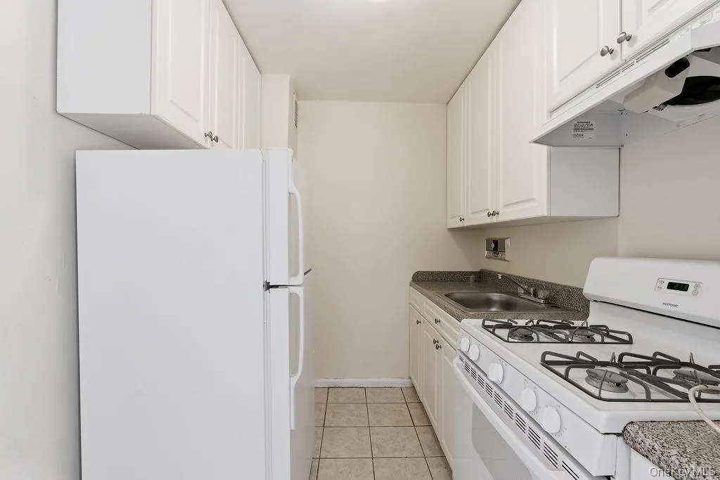 Kitchen featuring white appliances, under cabinet range hood, light tile patterned flooring, white cabinetry, and dark countertops Kitchen featuring white appliances, under cabinet range hood, light tile patterned flooring, white cabinetry, and dark countertops