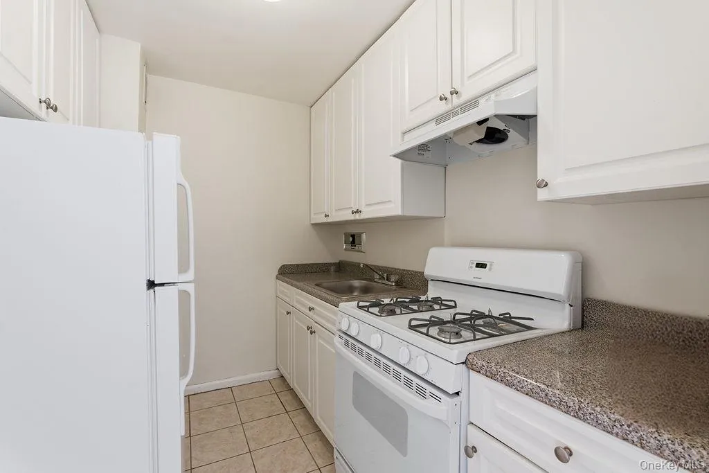Kitchen featuring white appliances, under cabinet range hood, light tile patterned floors, white cabinets, and dark countertops Kitchen featuring white appliances, under cabinet range hood, light tile patterned floors, white cabinets, and dark countertops