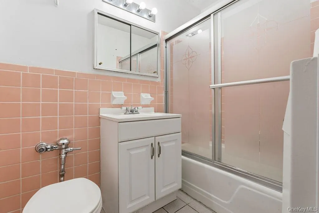 Full bathroom featuring tile walls, enclosed tub / shower combo, vanity, light tile patterned floors, and a wainscoted wall Full bathroom featuring tile walls, enclosed tub / shower combo, vanity, light tile patterned floors, and a wainscoted wall