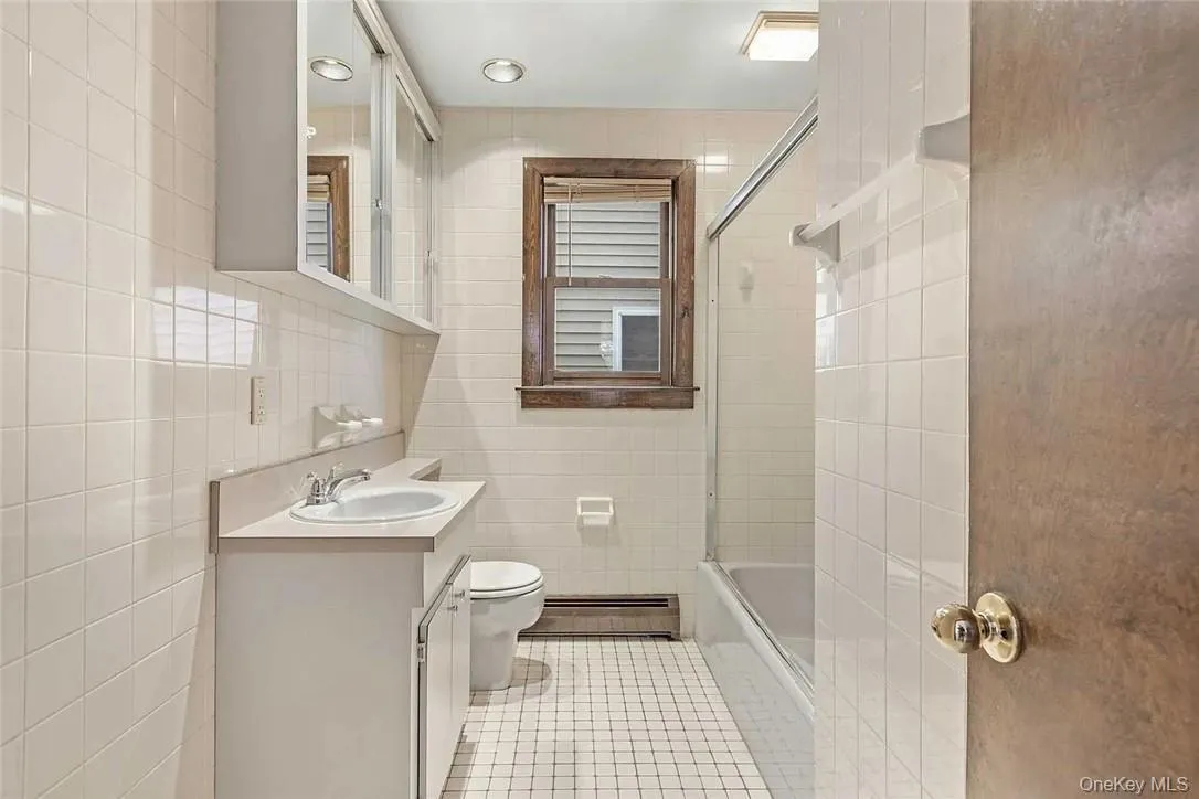 Bathroom featuring tile walls, vanity, light tile patterned floors, a baseboard radiator, and bath / shower combo with glass door Bathroom featuring tile walls, vanity, light tile patterned floors, a baseboard radiator, and bath / shower combo with glass door