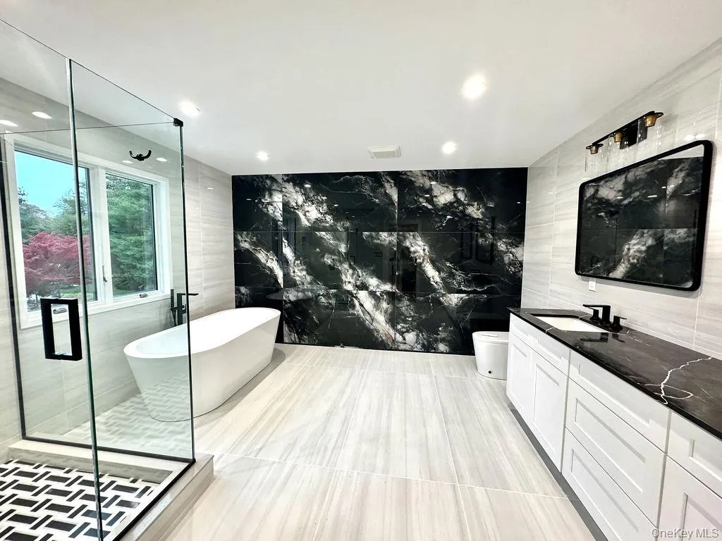 Bathroom featuring a freestanding soaking tub, a frameless glass shower enclosure with a patterned tile floor, a dual vanity with a dark stone countertop, a large accent wall with dark veined tile, and light-toned floor and wall tile Bathroom featuring a freestanding soaking tub, a frameless glass shower enclosure with a patterned tile floor, a dual vanity with a dark stone countertop, a large accent wall with dark veined tile, and light-toned floor and wall tile