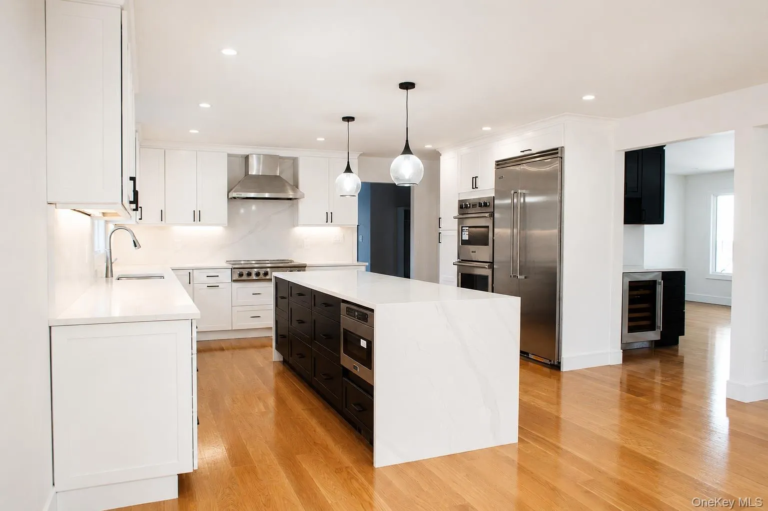 Open-concept kitchen featuring a waterfall island with integrated microwave, stainless steel appliances, white cabinetry with dark hardware, and wood-finish flooring Open-concept kitchen featuring a waterfall island with integrated microwave, stainless steel appliances, white cabinetry with dark hardware, and wood-finish flooring