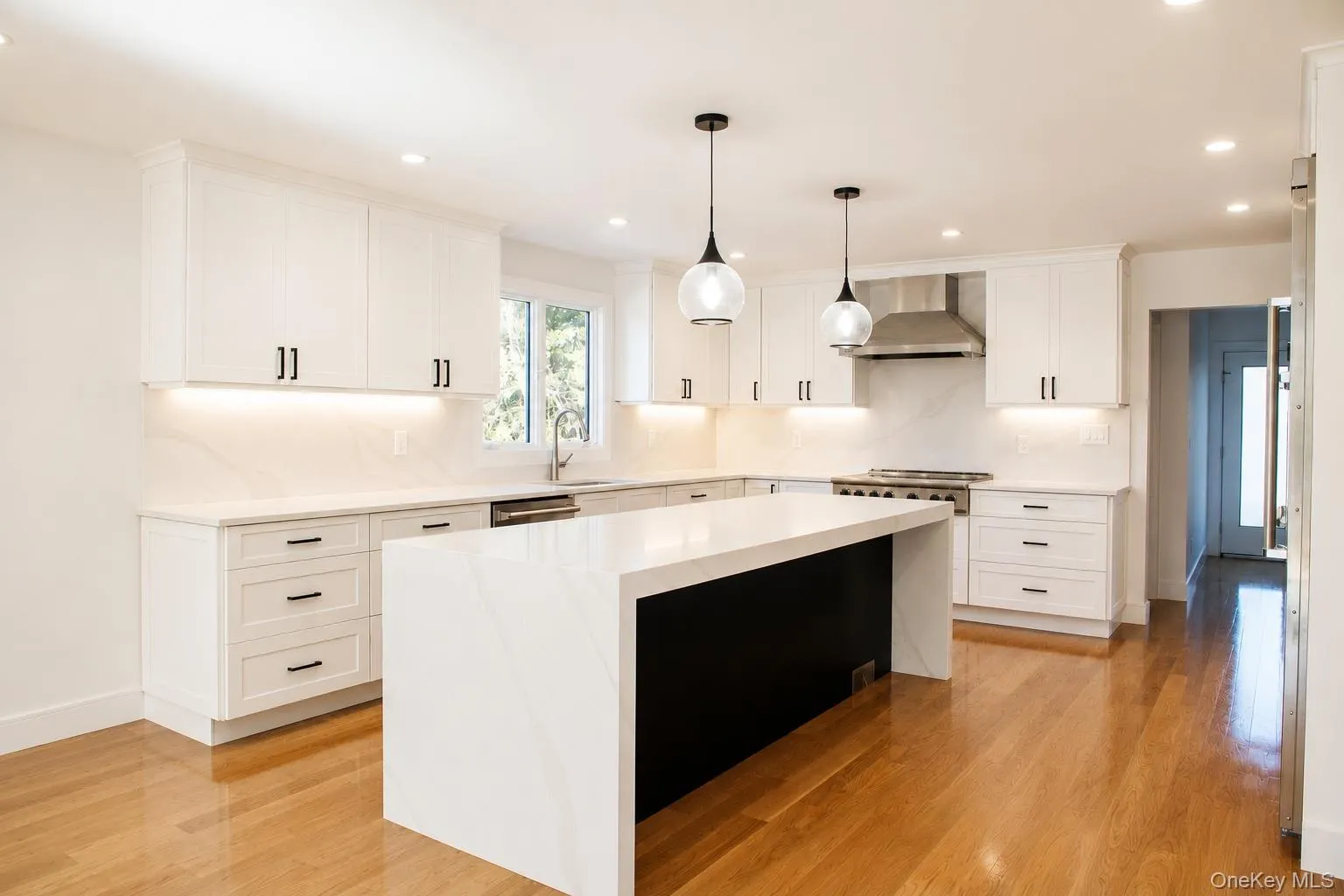 Kitchen featuring an expansive center island with waterfall countertop, white shaker cabinetry, stainless steel appliances, and wood-finish flooring Kitchen featuring an expansive center island with waterfall countertop, white shaker cabinetry, stainless steel appliances, and wood-finish flooring