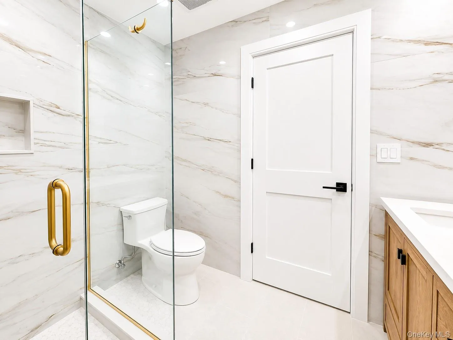 Bathroom featuring a glass-enclosed shower with gold-tone hardware, a white toilet, and white tile flooring Bathroom featuring a glass-enclosed shower with gold-tone hardware, a white toilet, and white tile flooring
