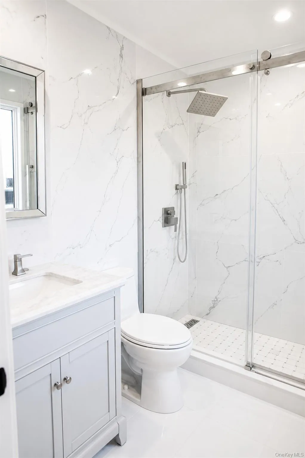 Contemporary bathroom featuring a frameless glass shower enclosure, a square rain showerhead, light gray vanity cabinet, and white stone-finish wall tiling Contemporary bathroom featuring a frameless glass shower enclosure, a square rain showerhead, light gray vanity cabinet, and white stone-finish wall tiling