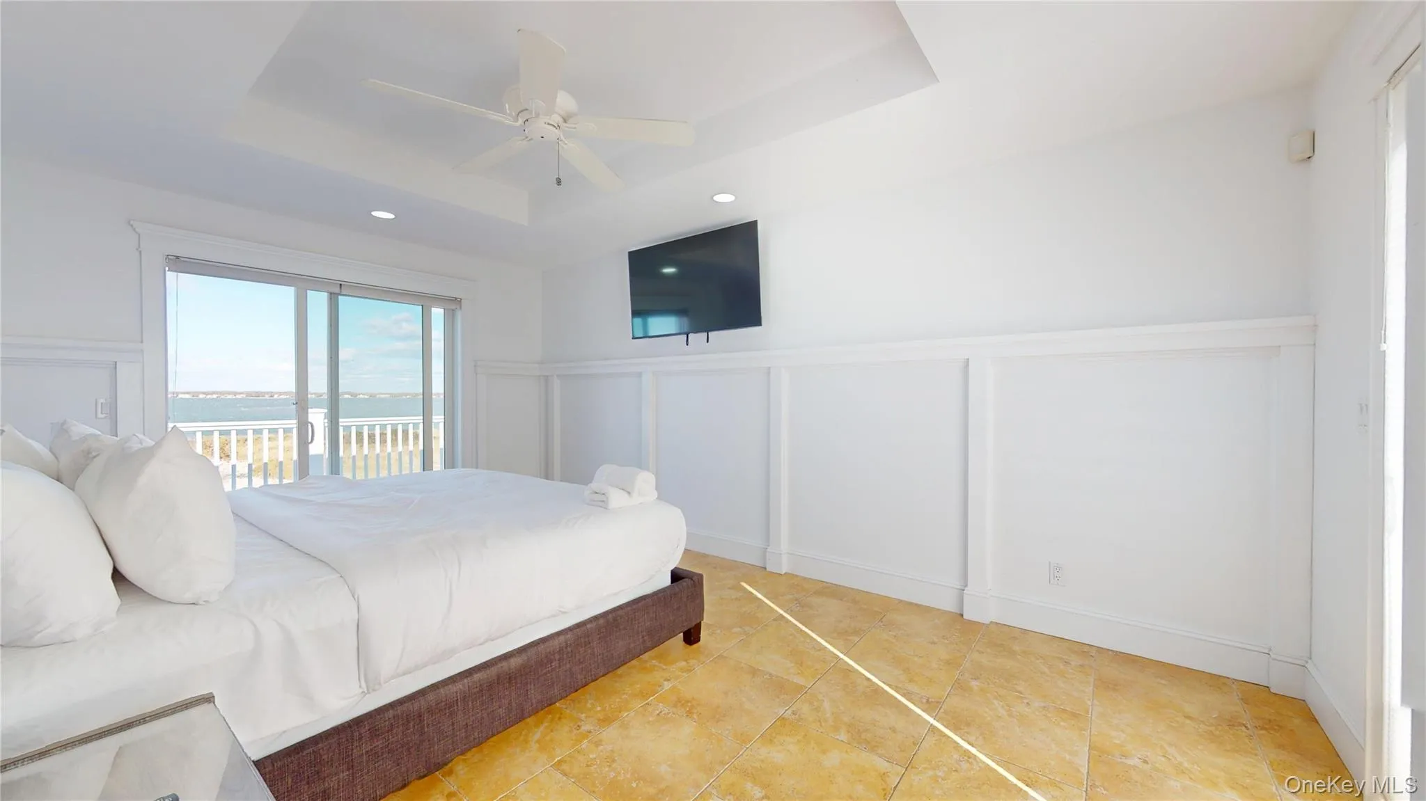 Bedroom featuring a tray ceiling, a decorative wall, ceiling fan, wainscoting, and access to exterior Bedroom featuring a tray ceiling, a decorative wall, ceiling fan, wainscoting, and access to exterior