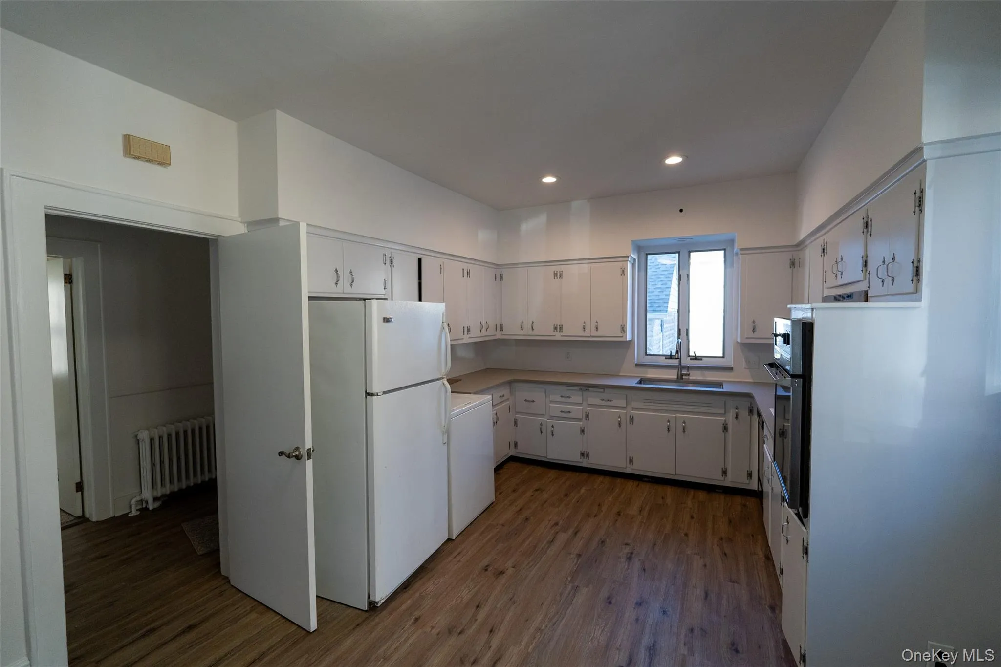 Kitchen with freestanding refrigerator, dark wood-style flooring, white cabinetry, light countertops, and radiator heating unit Kitchen with freestanding refrigerator, dark wood-style flooring, white cabinetry, light countertops, and radiator heating unit