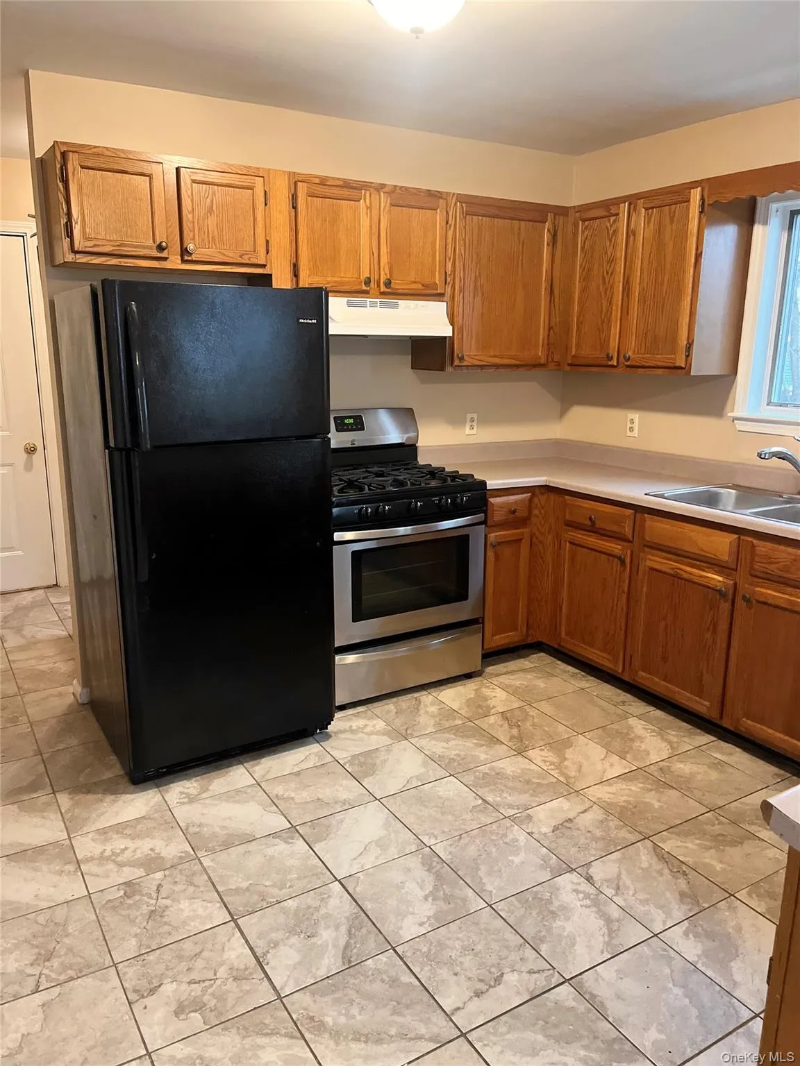 Kitchen featuring stainless steel gas stove, freestanding refrigerator, brown cabinetry, light countertops, and under cabinet range hood Kitchen featuring stainless steel gas stove, freestanding refrigerator, brown cabinetry, light countertops, and under cabinet range hood