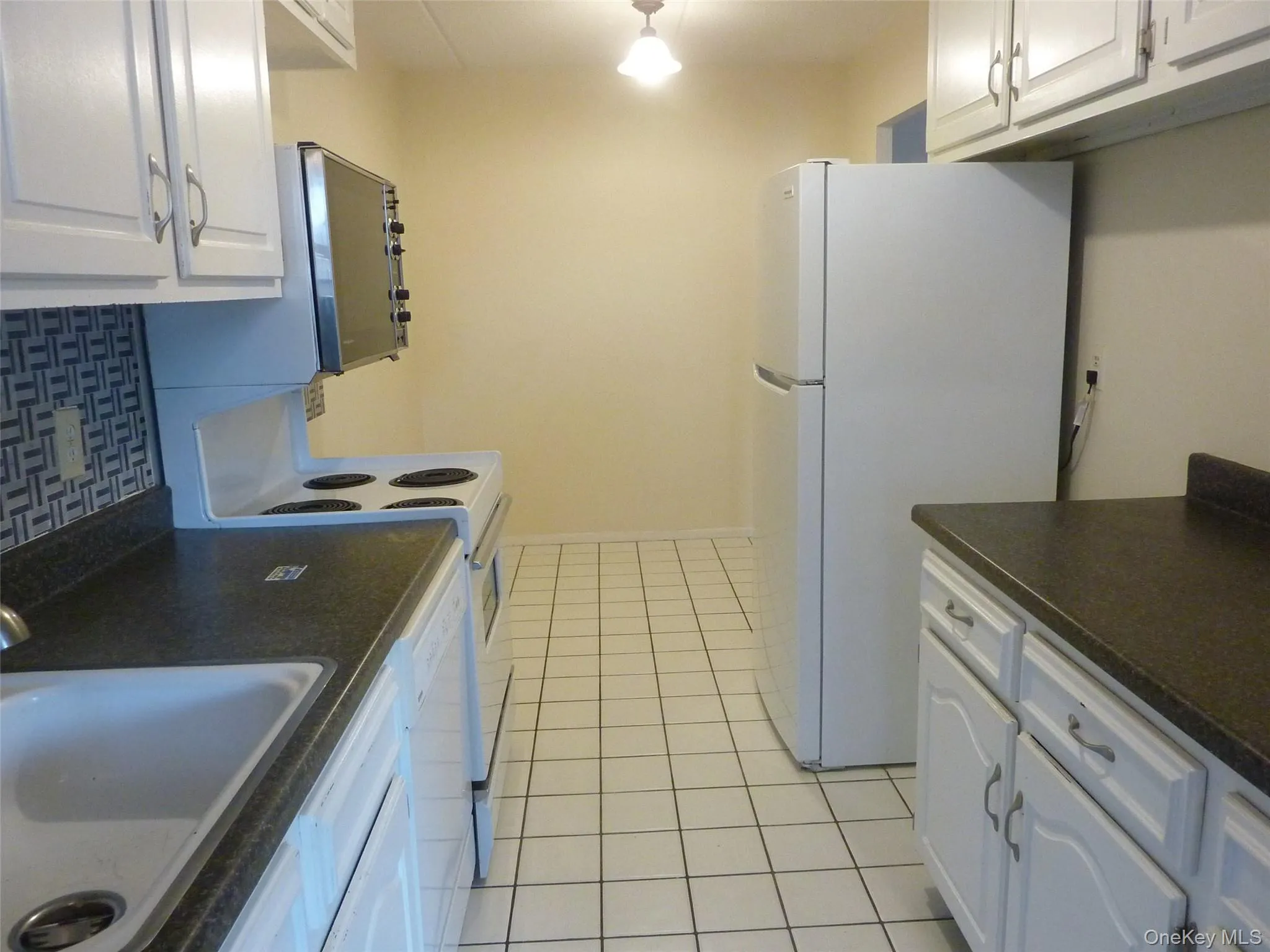 Kitchen with dark countertops, white cabinetry, white appliances, tasteful backsplash, and light tile patterned flooring Kitchen with dark countertops, white cabinetry, white appliances, tasteful backsplash, and light tile patterned flooring