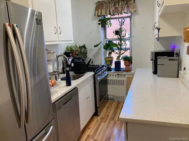 Kitchen featuring appliances with stainless steel finishes, white cabinetry, radiator, light wood-style floors, and light stone countertops Kitchen featuring appliances with stainless steel finishes, white cabinetry, radiator, light wood-style floors, and light stone countertops