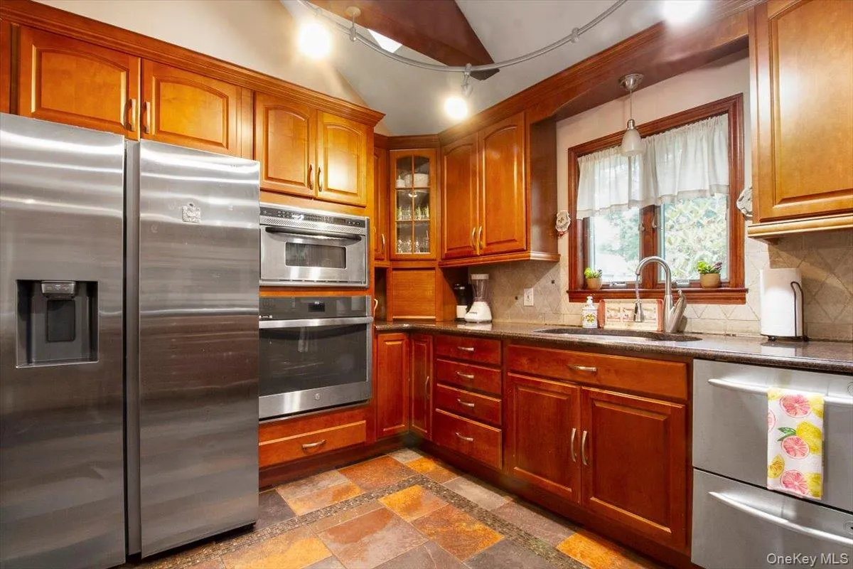Kitchen with appliances with stainless steel finishes, glass insert cabinets, dark stone counters, decorative backsplash, and brown cabinetry Kitchen with appliances with stainless steel finishes, glass insert cabinets, dark stone counters, decorative backsplash, and brown cabinetry