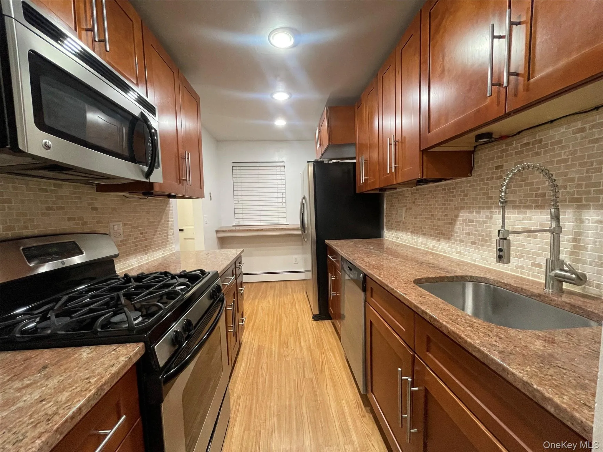 Kitchen featuring appliances with stainless steel finishes, brown cabinets, light stone counters, light wood-type flooring, and recessed lighting Kitchen featuring appliances with stainless steel finishes, brown cabinets, light stone counters, light wood-type flooring, and recessed lighting
