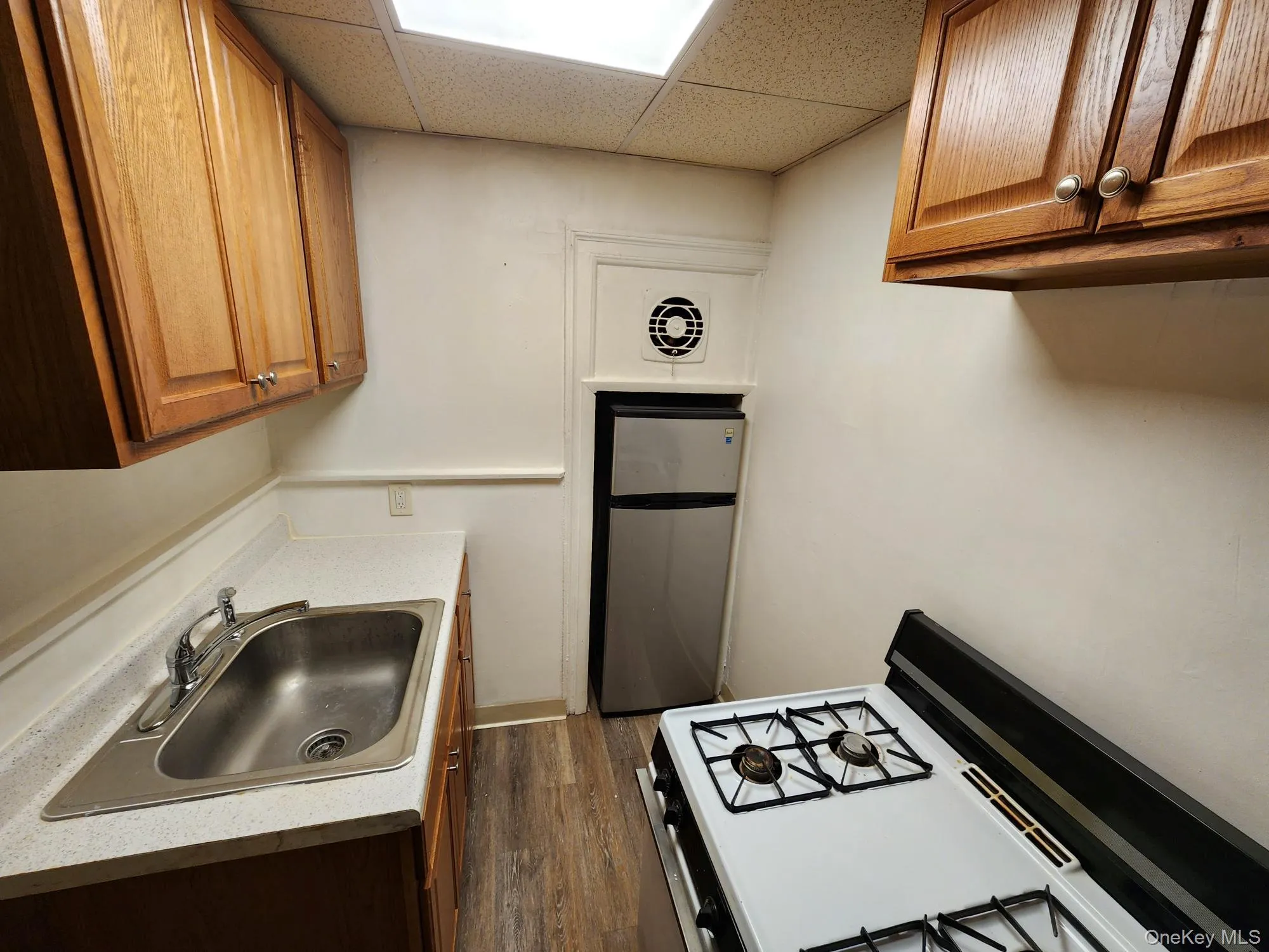 Kitchen featuring brown cabinetry, a drop ceiling, light countertops, dark wood-style flooring, and white range with gas cooktop Kitchen featuring brown cabinetry, a drop ceiling, light countertops, dark wood-style flooring, and white range with gas cooktop