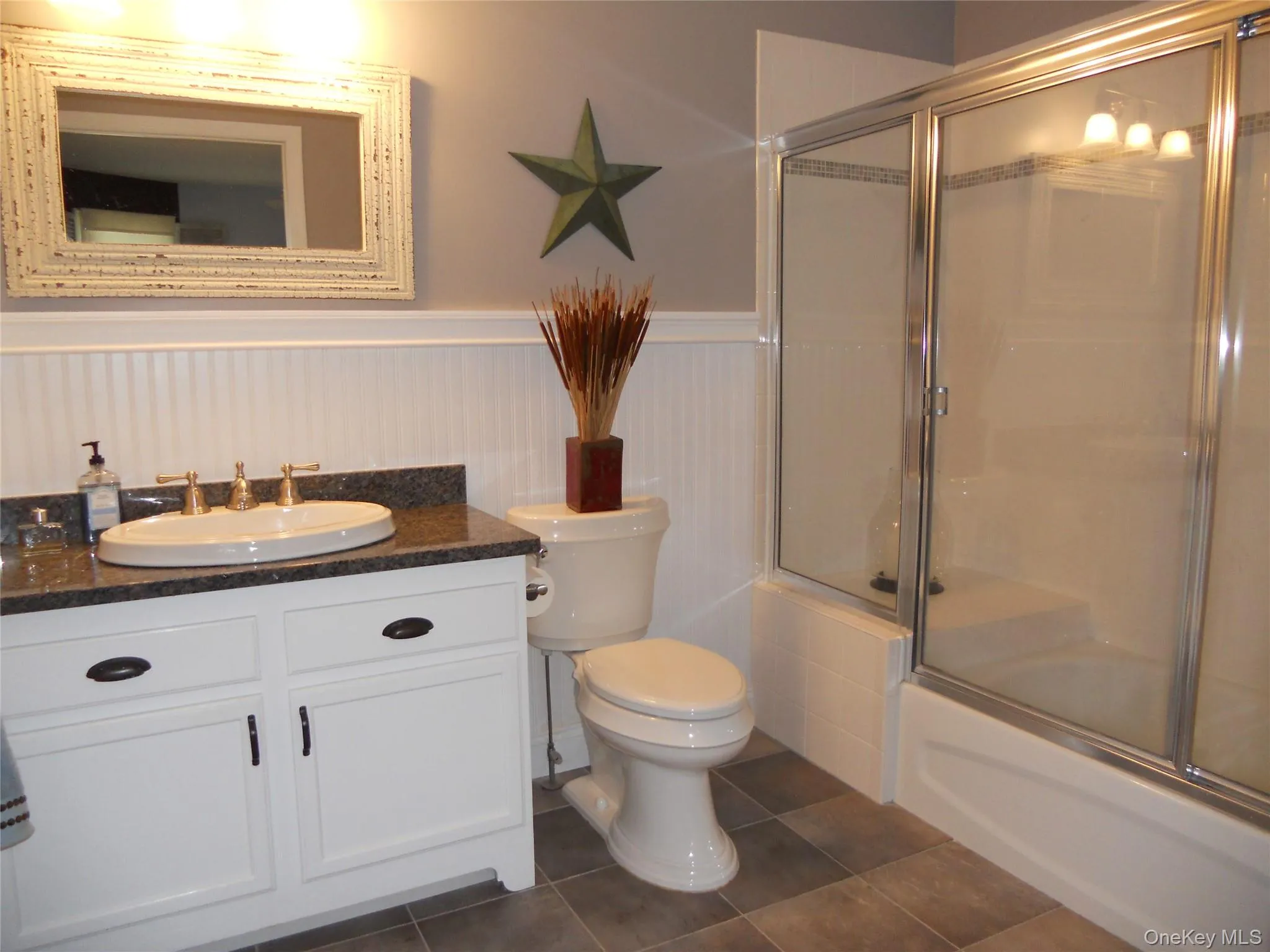 Full bathroom with vanity, a wainscoted wall, shower / bath combination with glass door, and dark tile patterned floors Full bathroom with vanity, a wainscoted wall, shower / bath combination with glass door, and dark tile patterned floors