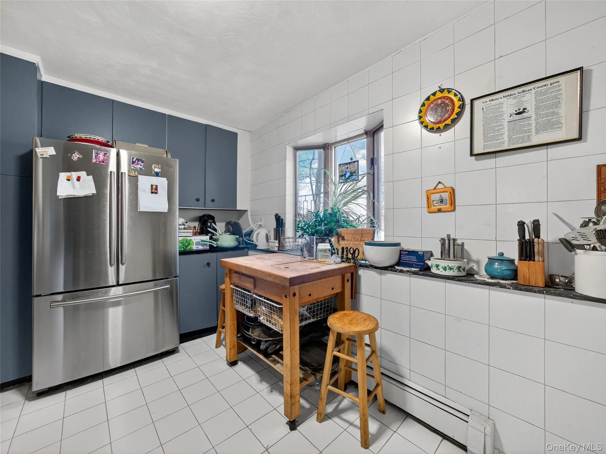 Kitchen with dark countertops, freestanding refrigerator, a baseboard radiator, blue cabinetry, and light tile patterned floors Kitchen with dark countertops, freestanding refrigerator, a baseboard radiator, blue cabinetry, and light tile patterned floors