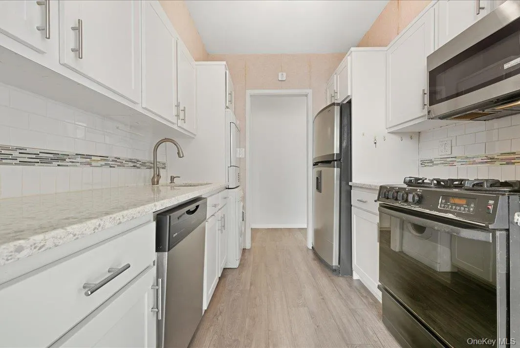 Kitchen with stainless steel appliances, backsplash, white cabinets, light wood-type flooring, and light stone counters Kitchen with stainless steel appliances, backsplash, white cabinets, light wood-type flooring, and light stone counters