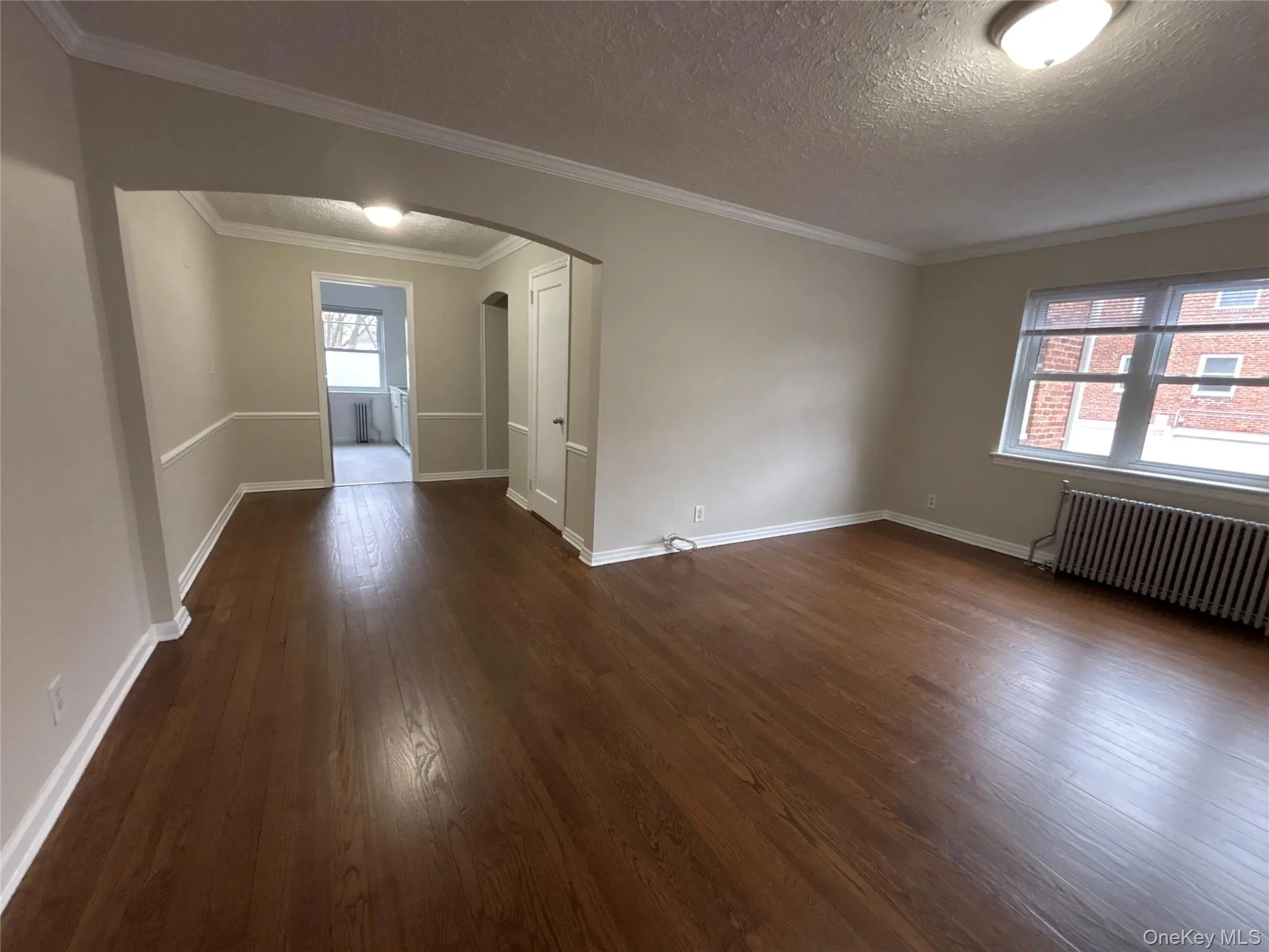 Empty room featuring arched walkways, radiator, dark wood-type flooring, crown molding, and a textured ceiling Empty room featuring arched walkways, radiator, dark wood-type flooring, crown molding, and a textured ceiling