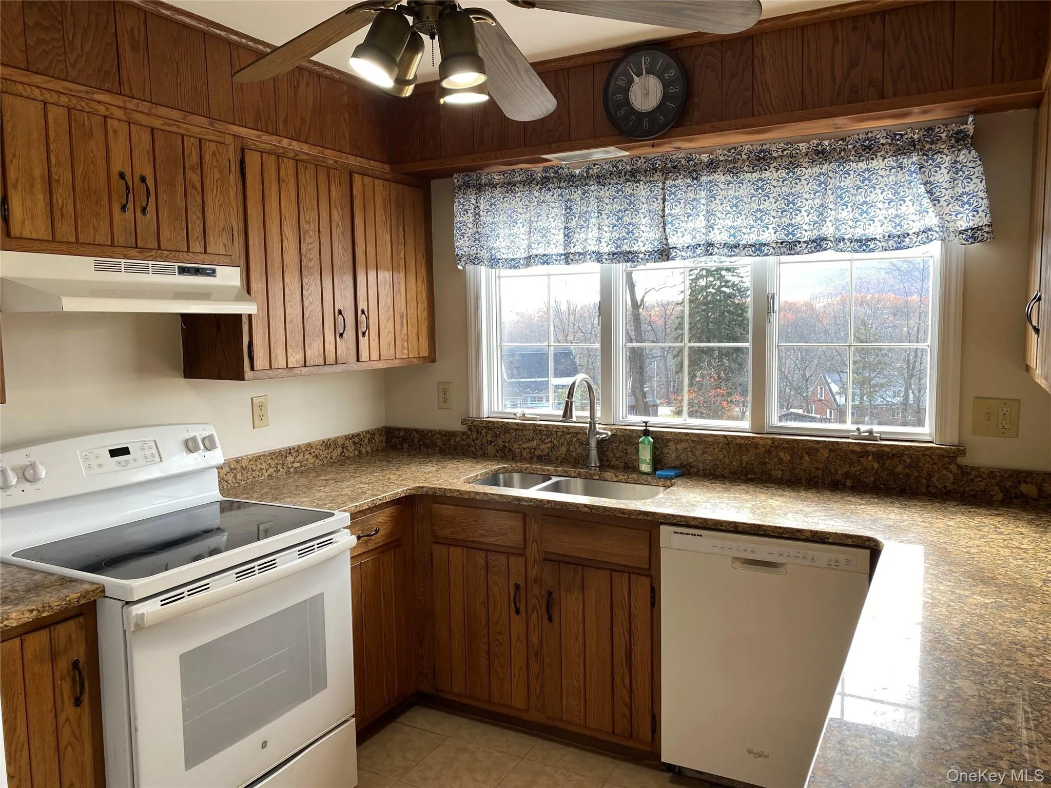 Kitchen with white appliances, under cabinet range hood, a ceiling fan, brown cabinetry, and dark countertops Kitchen with white appliances, under cabinet range hood, a ceiling fan, brown cabinetry, and dark countertops