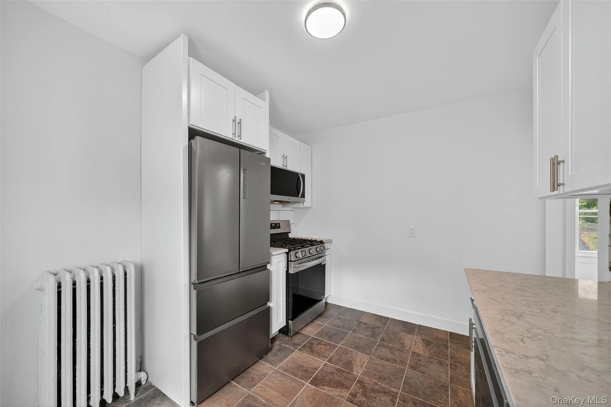 Kitchen featuring radiator heating unit, stainless steel appliances, light countertops, white cabinetry, and stone finish floors Kitchen featuring radiator heating unit, stainless steel appliances, light countertops, white cabinetry, and stone finish floors