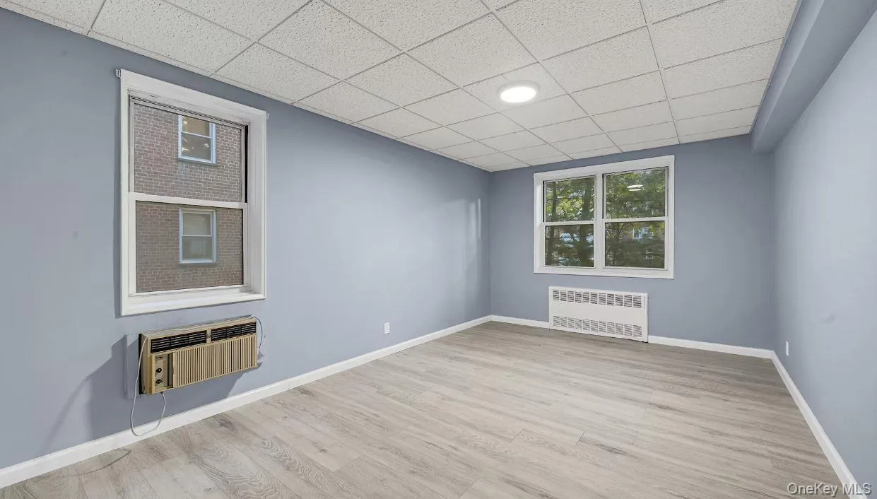 Bedroom featuring two windows, wood finished floors, radiator heating unit, a drop ceiling, and a wall unit AC Bedroom featuring two windows, wood finished floors, radiator heating unit, a drop ceiling, and a wall unit AC