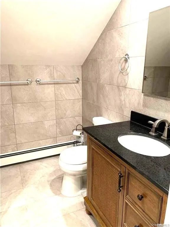 Bathroom with tile walls, a baseboard radiator, lofted ceiling, vanity, and light tile patterned floors Bathroom with tile walls, a baseboard radiator, lofted ceiling, vanity, and light tile patterned floors