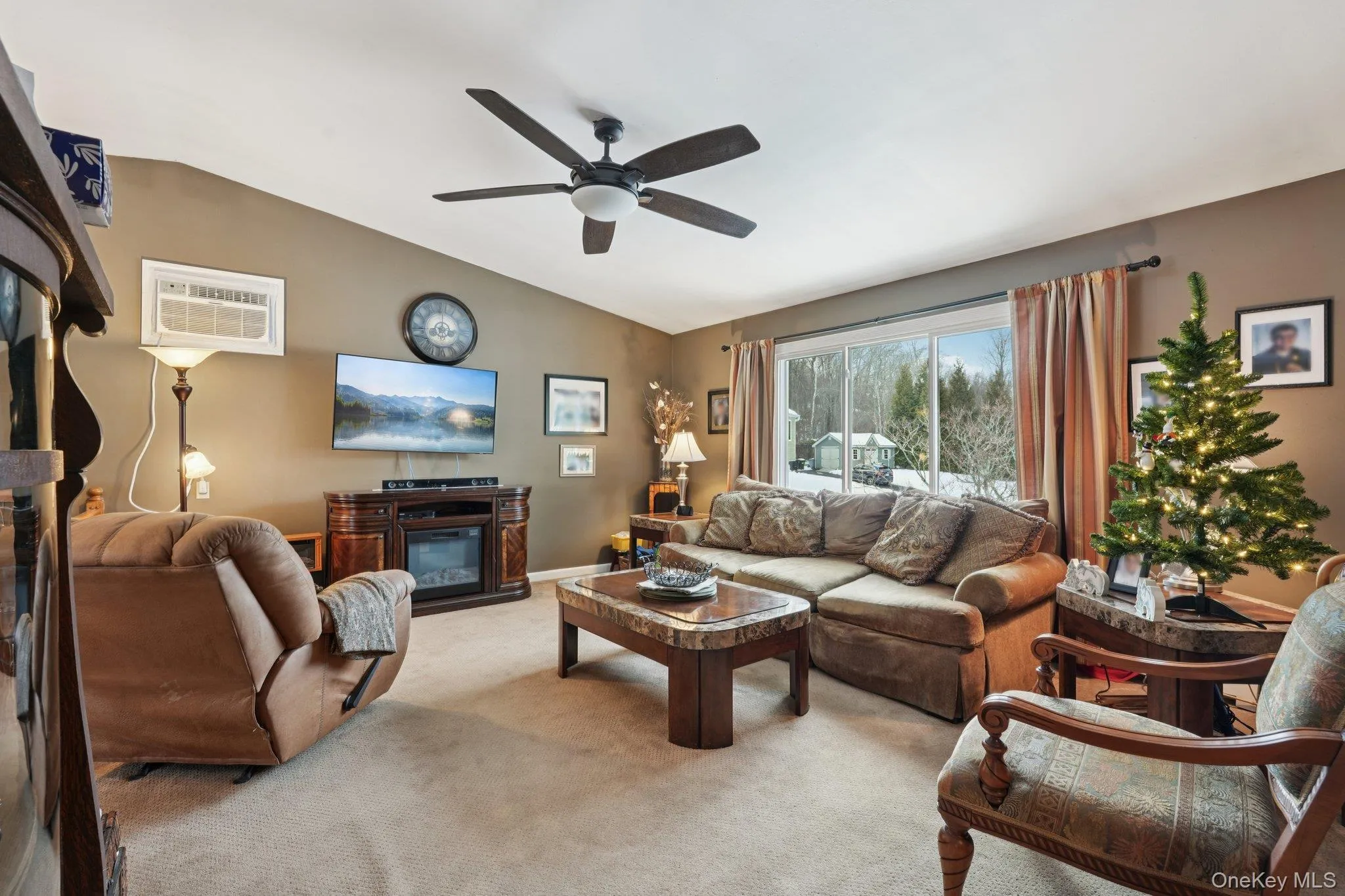 Carpeted living area with a glass covered fireplace, vaulted ceiling, ceiling fan, and a wall mounted AC. beautiful in all seasons. Digitally enhanced photo Carpeted living area with a glass covered fireplace, vaulted ceiling, ceiling fan, and a wall mounted AC. beautiful in all seasons. Digitally enhanced photo