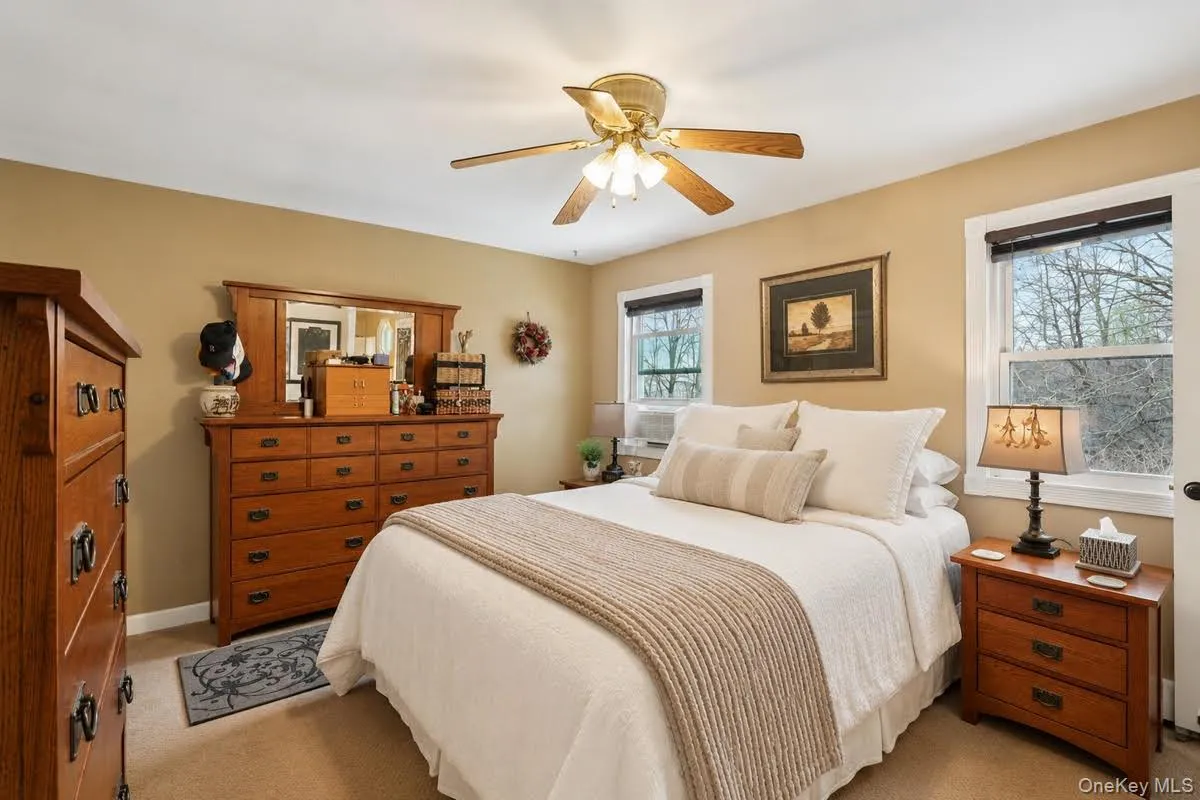 Carpeted sleeping area featuring a ceiling fan, two windows with white trim, and a built-in air conditioning unit. some photos may include virtually staged or digitally modified Carpeted sleeping area featuring a ceiling fan, two windows with white trim, and a built-in air conditioning unit. some photos may include virtually staged or digitally modified