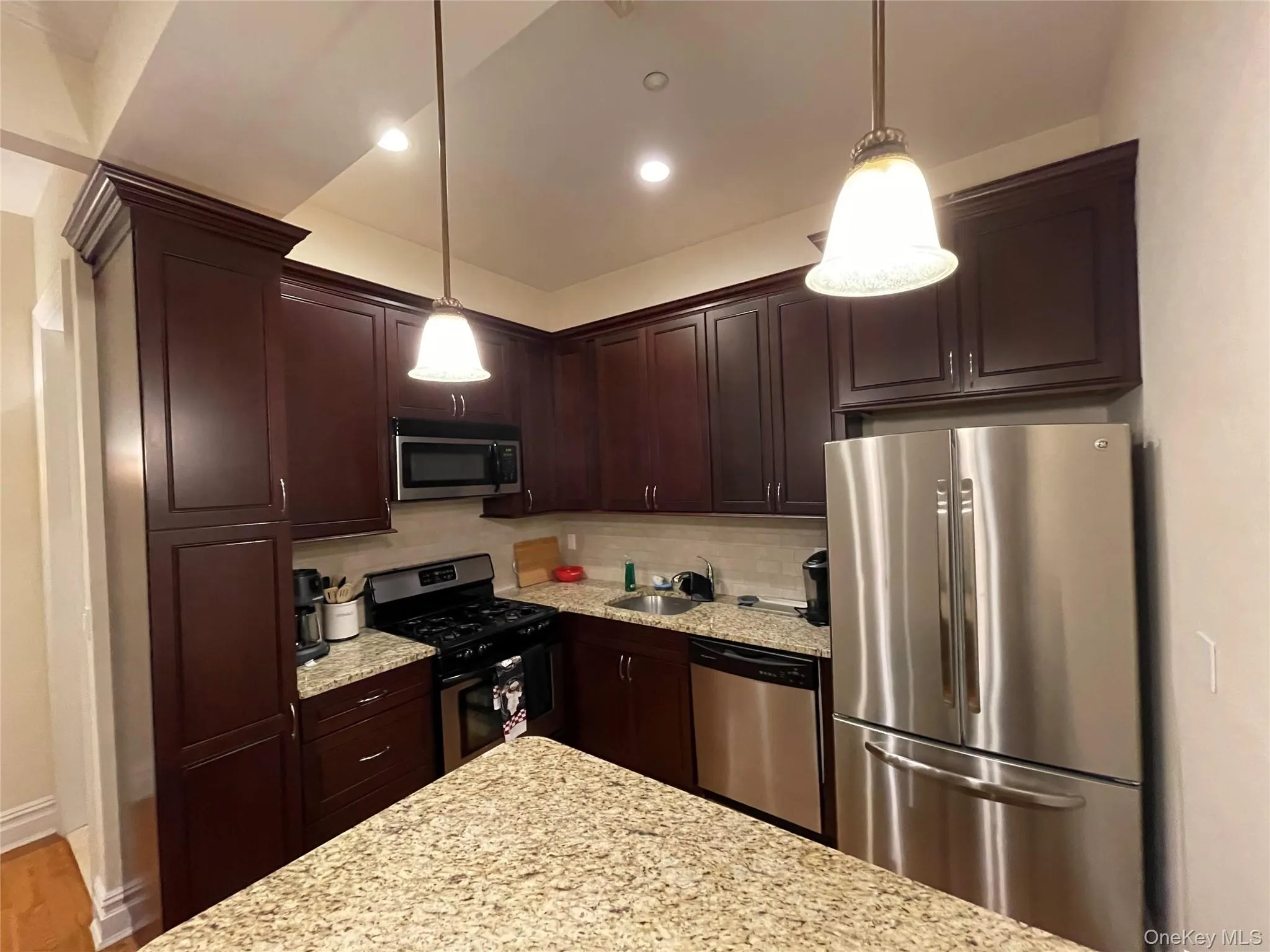 Kitchen featuring stainless steel appliances, light stone counters, tasteful backsplash, dark brown cabinets, and decorative light fixtures Kitchen featuring stainless steel appliances, light stone counters, tasteful backsplash, dark brown cabinets, and decorative light fixtures
