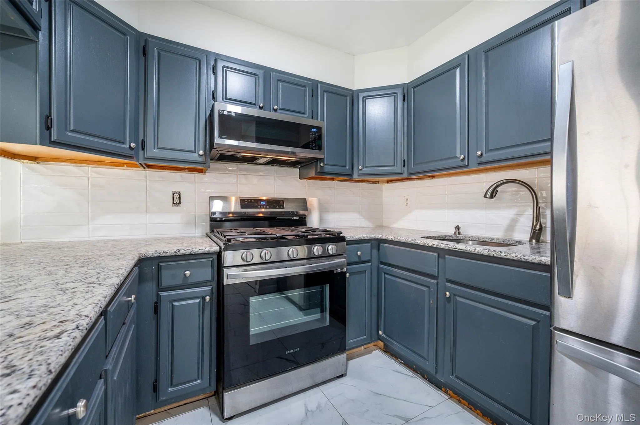 Kitchen featuring appliances with stainless steel finishes, backsplash, light stone counters, and blue cabinetry Kitchen featuring appliances with stainless steel finishes, backsplash, light stone counters, and blue cabinetry