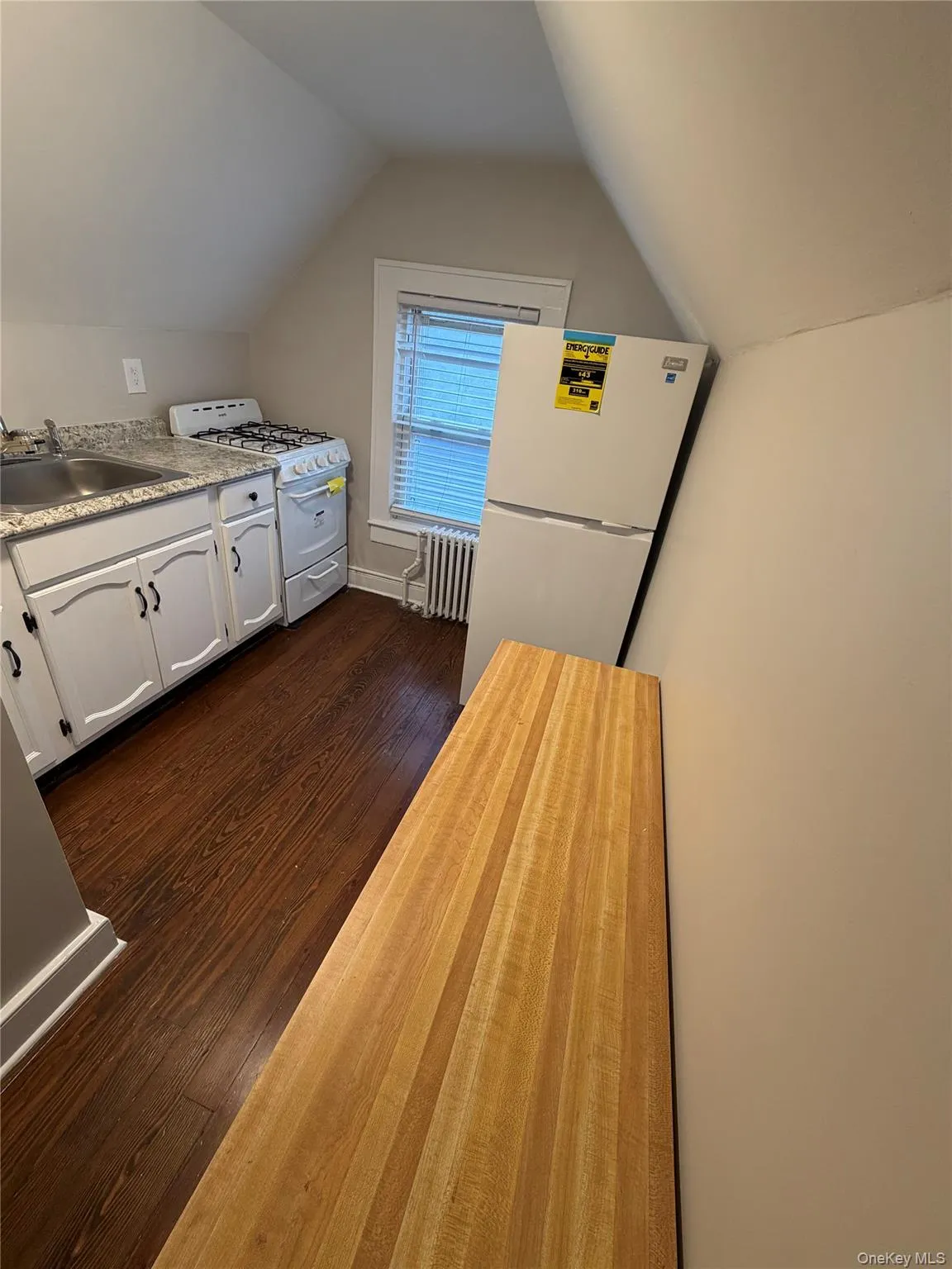 Kitchen with white appliances, lofted ceiling, dark wood-style floors, white cabinets, and radiator heating unit Kitchen with white appliances, lofted ceiling, dark wood-style floors, white cabinets, and radiator heating unit