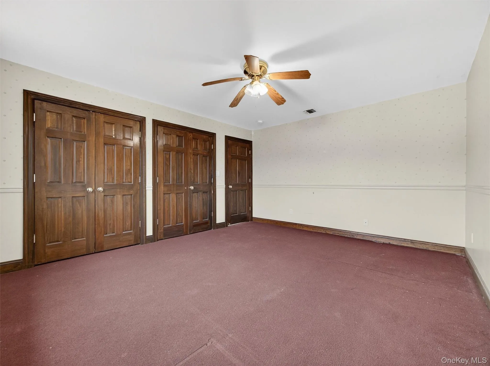 Unfurnished bedroom featuring multiple closets, wallpapered walls, ceiling fan, and carpet floors Unfurnished bedroom featuring multiple closets, wallpapered walls, ceiling fan, and carpet floors