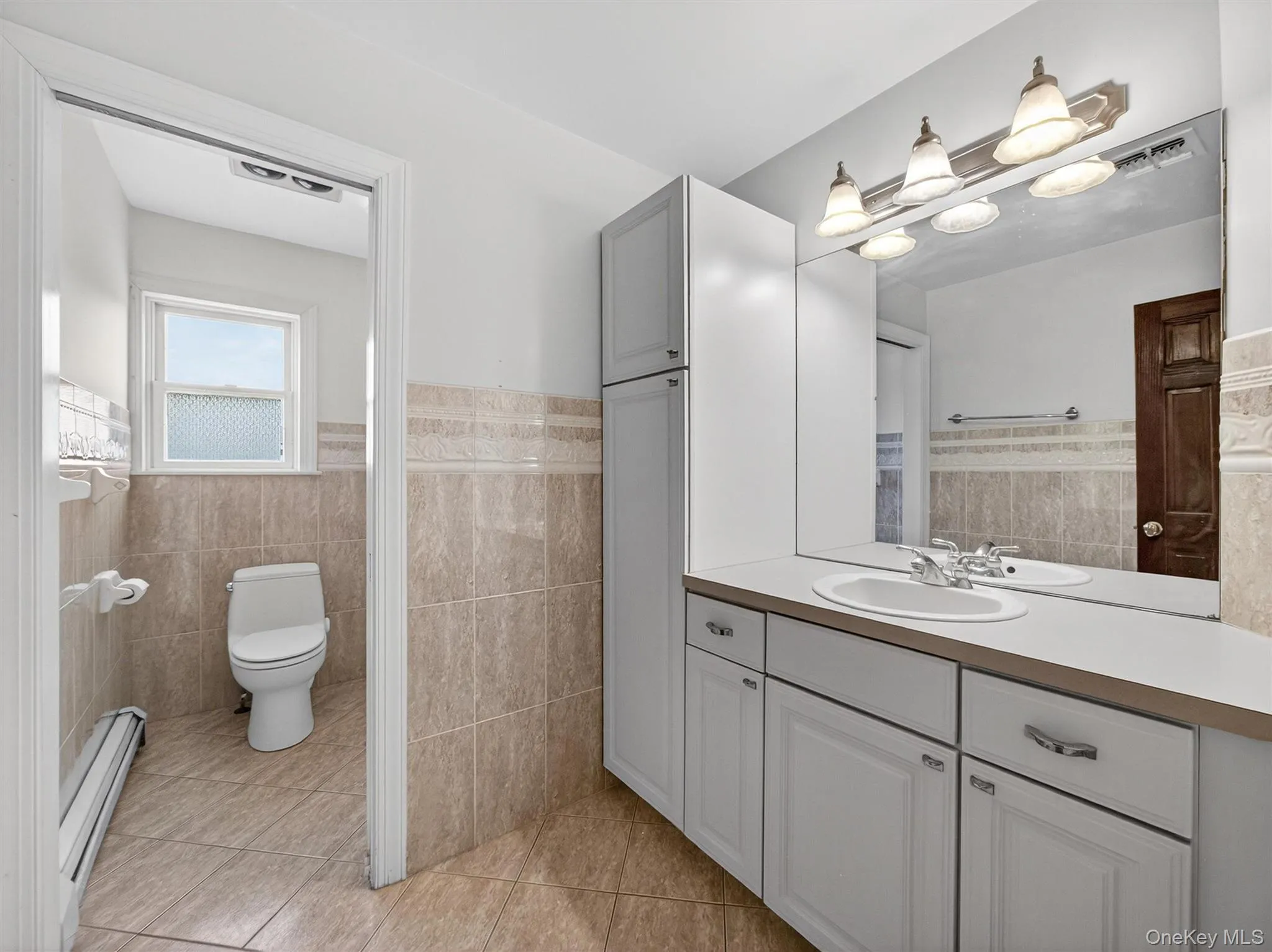 Bathroom with a wainscoted wall, tile walls, vanity, a baseboard radiator, and light tile patterned floors Bathroom with a wainscoted wall, tile walls, vanity, a baseboard radiator, and light tile patterned floors