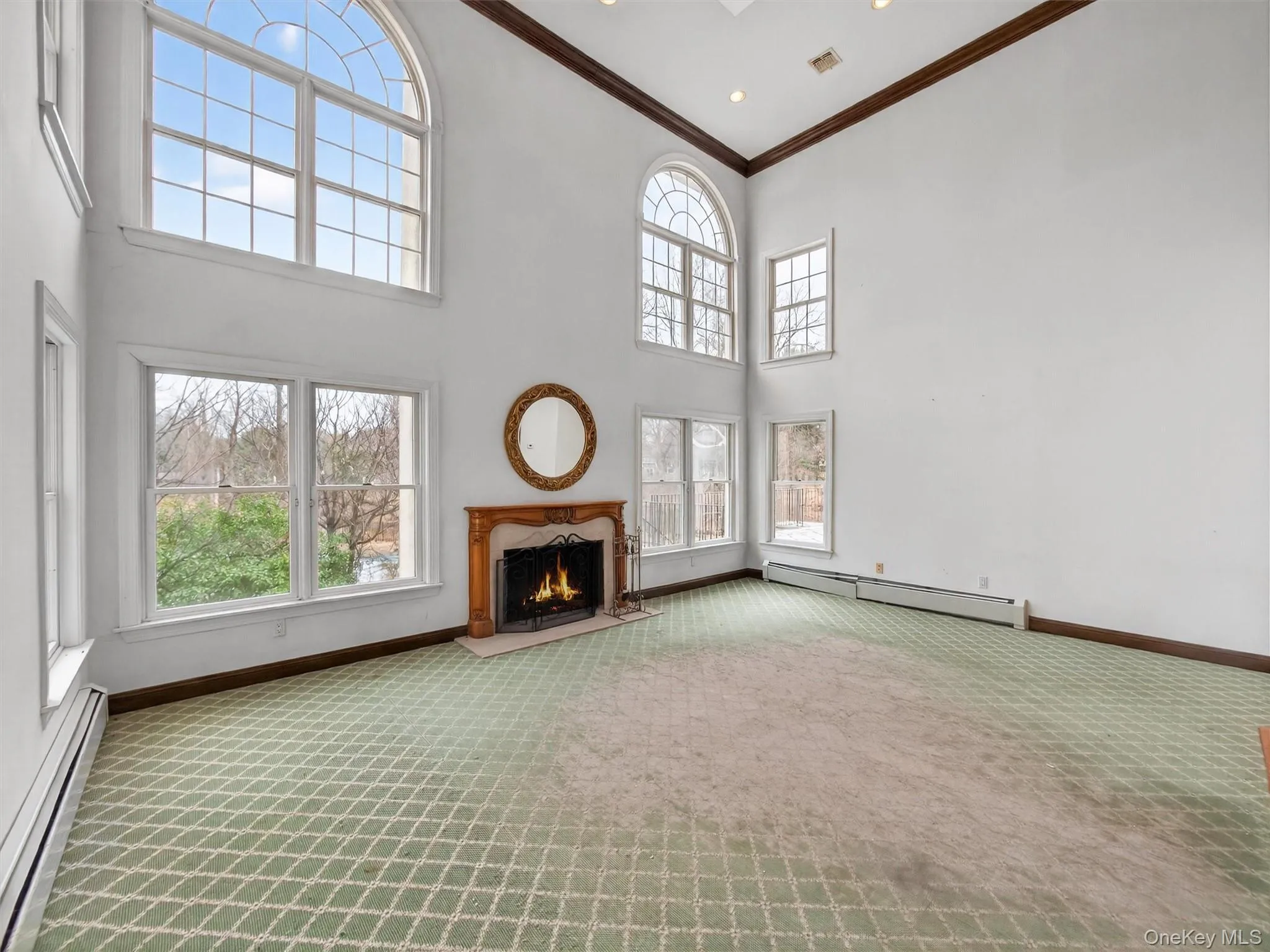 Unfurnished living room with baseboard heating, a warm lit fireplace, light carpet, a high ceiling, and crown molding Unfurnished living room with baseboard heating, a warm lit fireplace, light carpet, a high ceiling, and crown molding