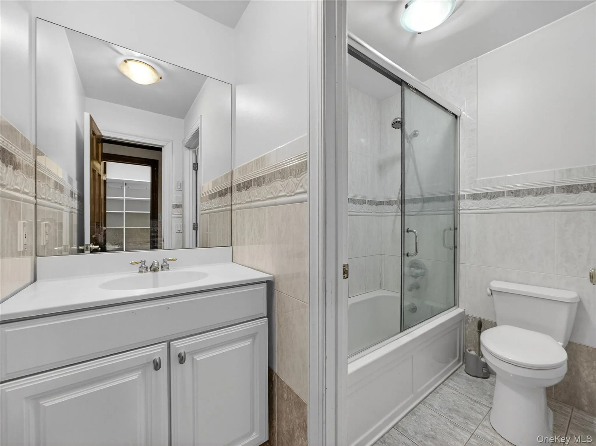 Full bathroom with tile walls, vanity, bath / shower combo with glass door, wainscoting, and light tile patterned flooring Full bathroom with tile walls, vanity, bath / shower combo with glass door, wainscoting, and light tile patterned flooring