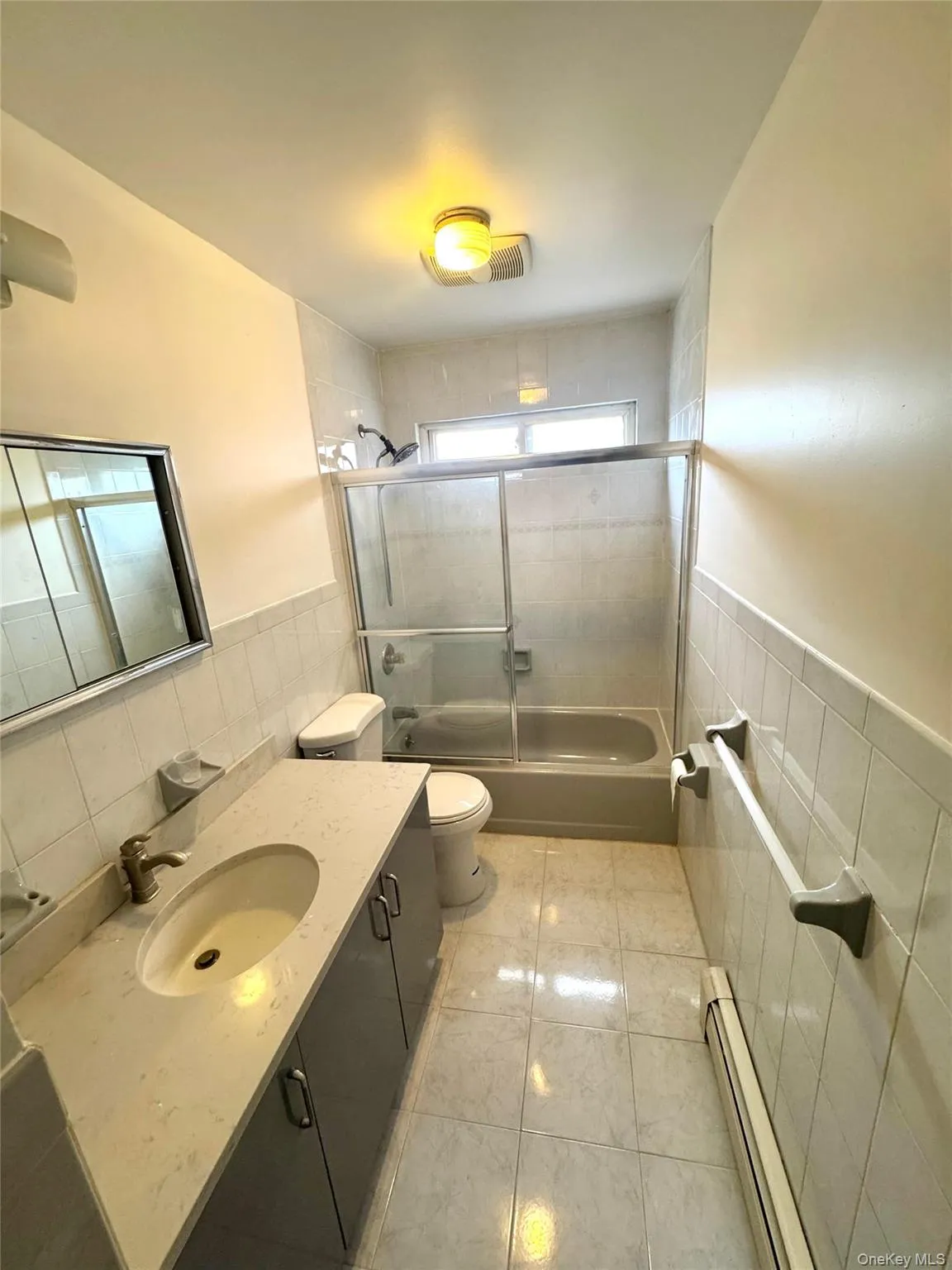 Bathroom featuring tile walls, a wainscoted wall, vanity, a baseboard heating unit, and bath / shower combo with glass door Bathroom featuring tile walls, a wainscoted wall, vanity, a baseboard heating unit, and bath / shower combo with glass door